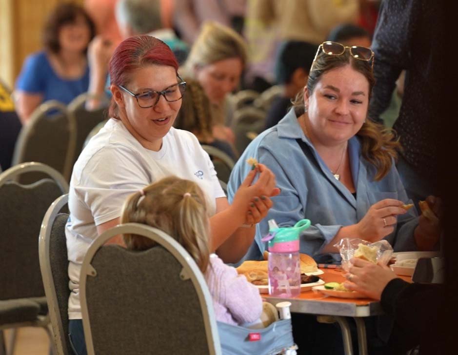 People sitting at a table, eating and socializing in a busy indoor setting, possibly a community gathering or event.