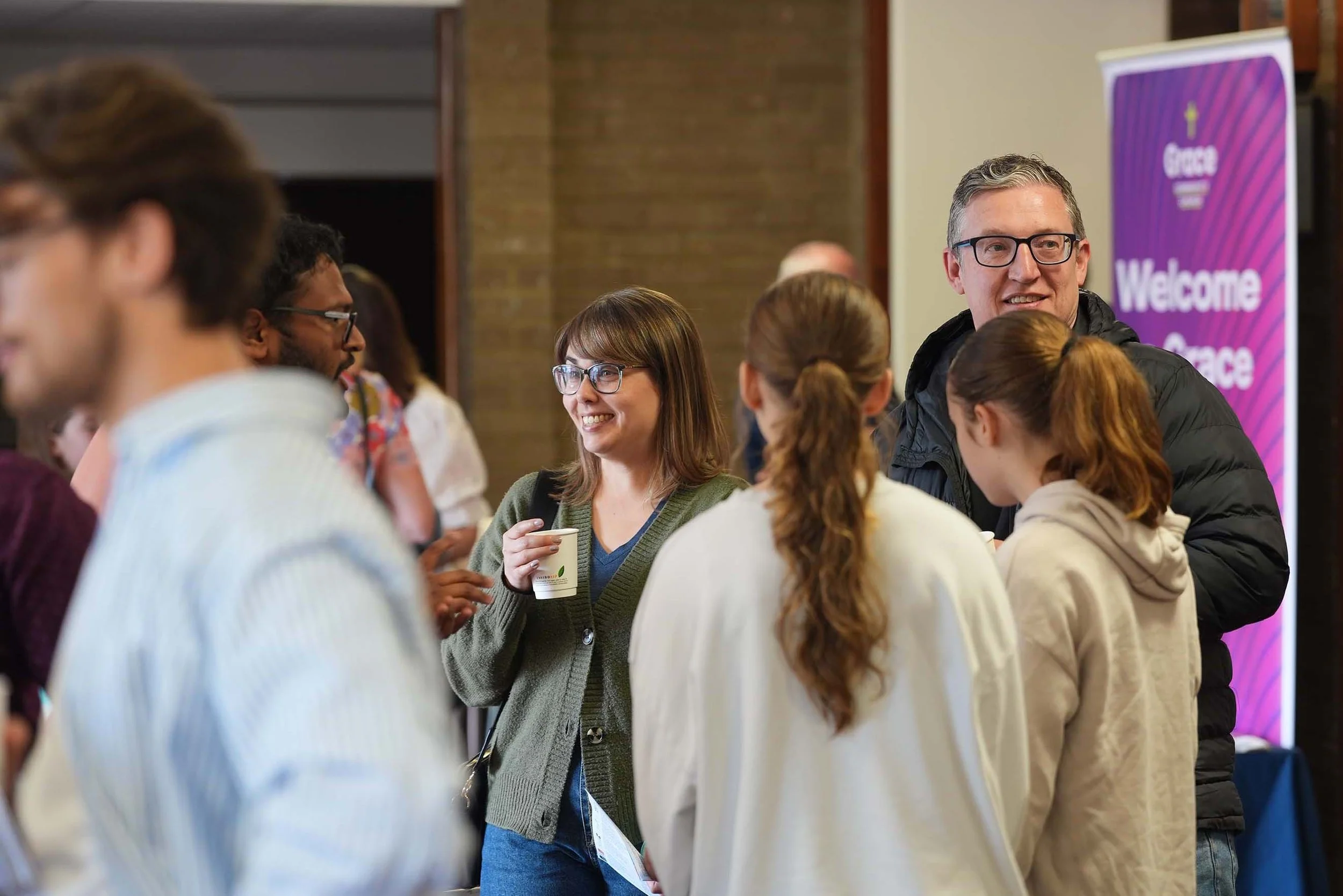 People socializing and chatting at an indoor event, with a purple sign in the background that reads 'Grace Welcome'.