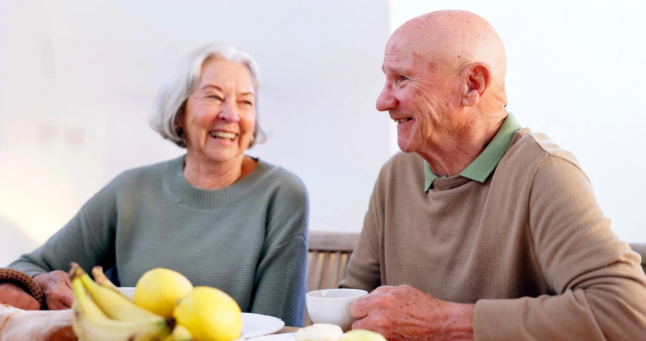 An elderly woman and man smiling and talking at a table with bananas, lemons, and baked goods.