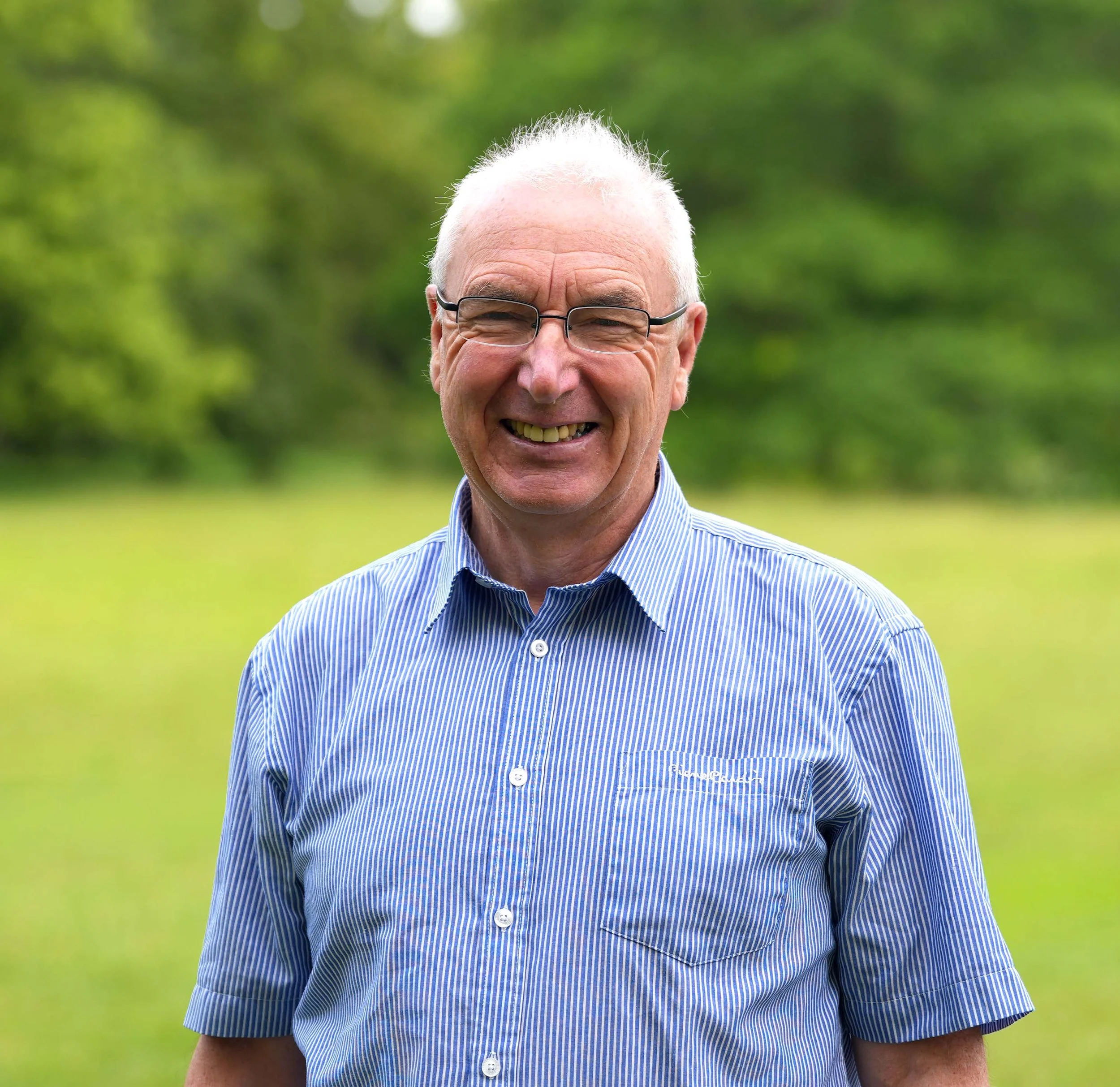 A smiling elderly man wearing glasses and a blue and white striped collared shirt outdoors with green trees and grass in the background.