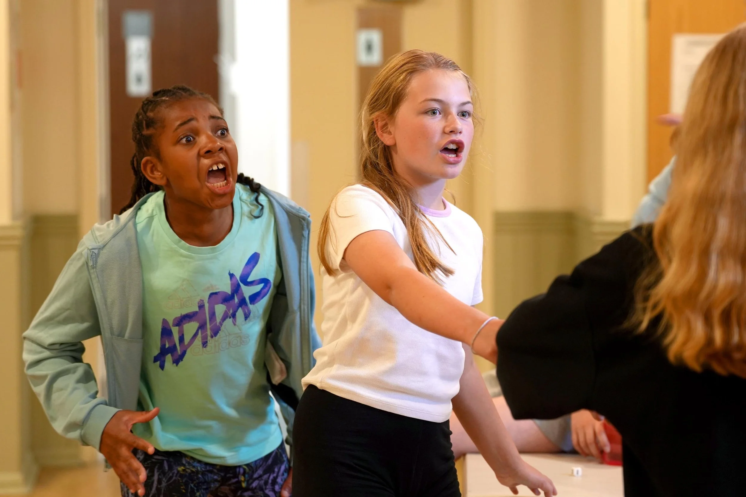 Two girls standing in line, one with a surprised expression and the other looking forward, while a person with long hair is reaching across the table in the foreground. The setting appears to be indoors, possibly a school or community center.