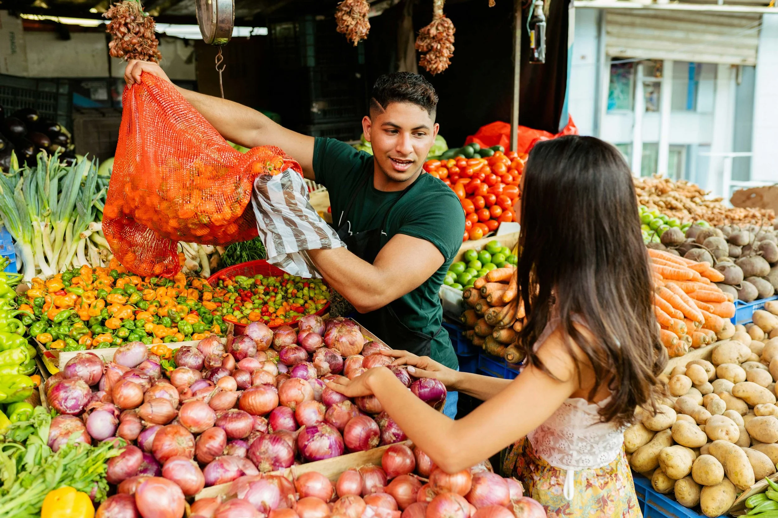 A young man is selling fresh vegetables, including onions, carrots, and potatoes, at a market stall. He is engaging with a customer, a woman with long dark hair, while standing behind the produce. Bright natural light illuminates the vibrant display of colorful vegetables.