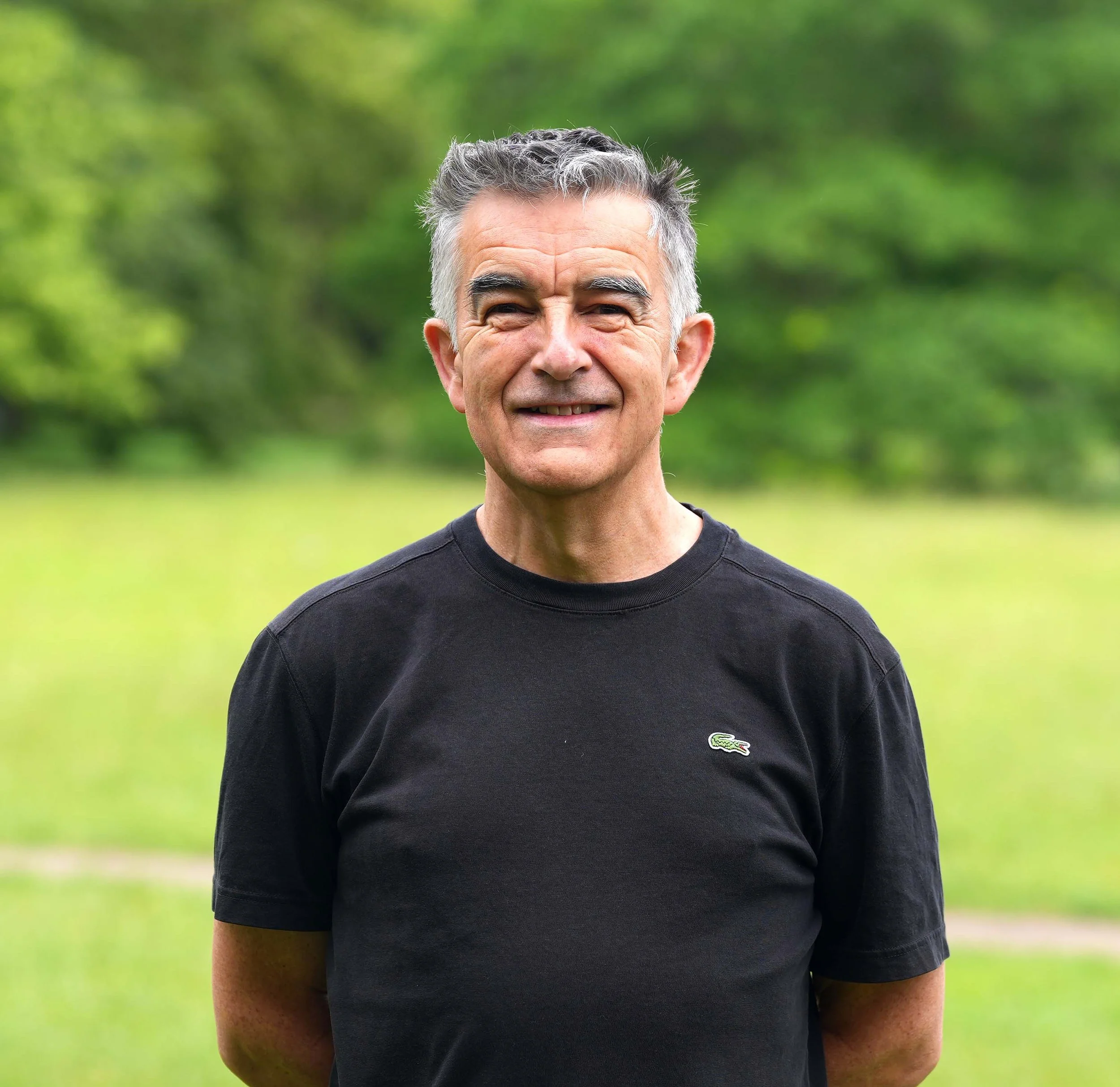 Close-up of a smiling middle-aged man with gray hair, wearing a black Lacoste T-shirt, standing outdoors with greenery in the background.