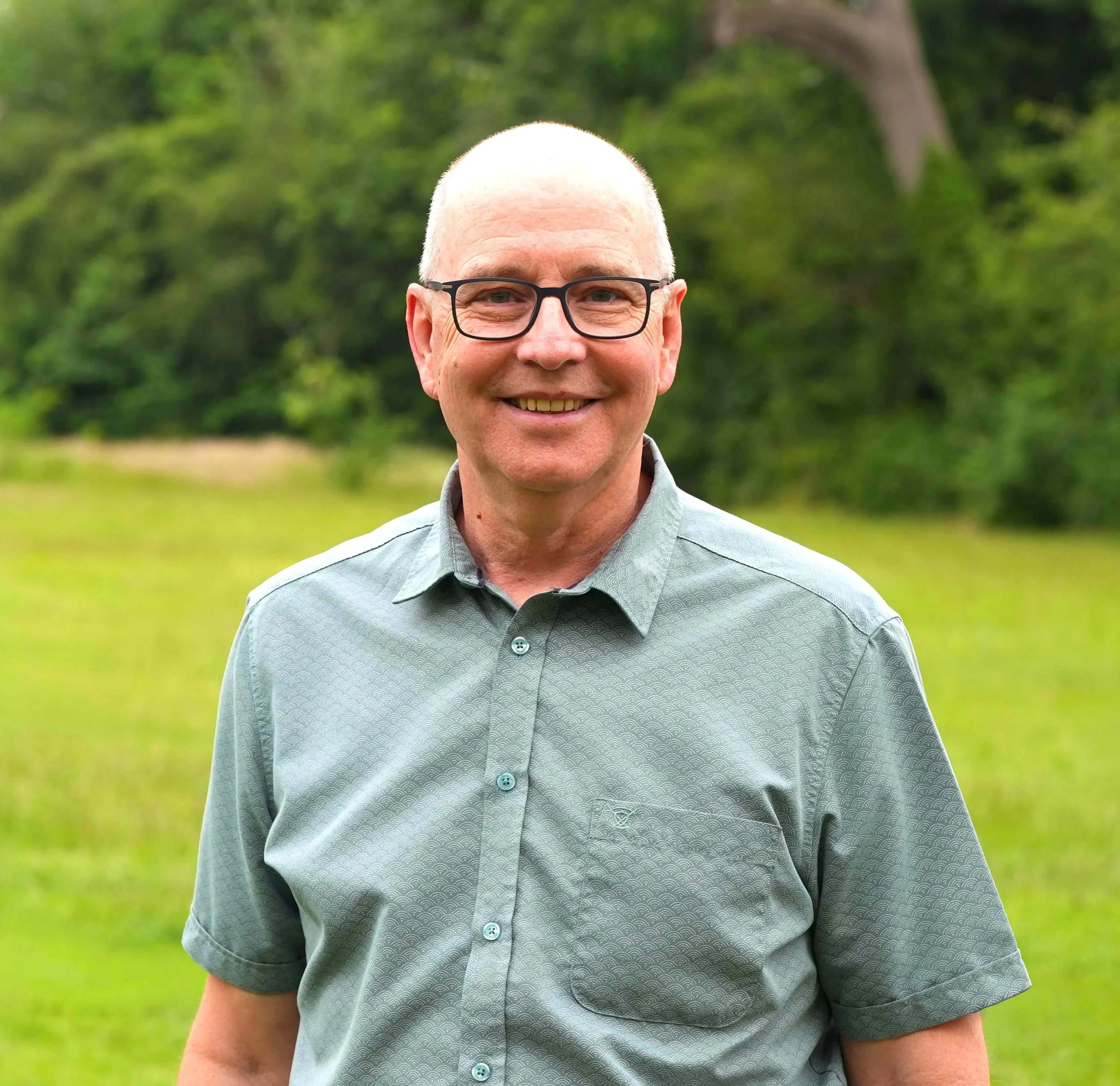 Close-up of a smiling middle-aged man with glasses and a patterned shirt outdoors, with green trees in the background.