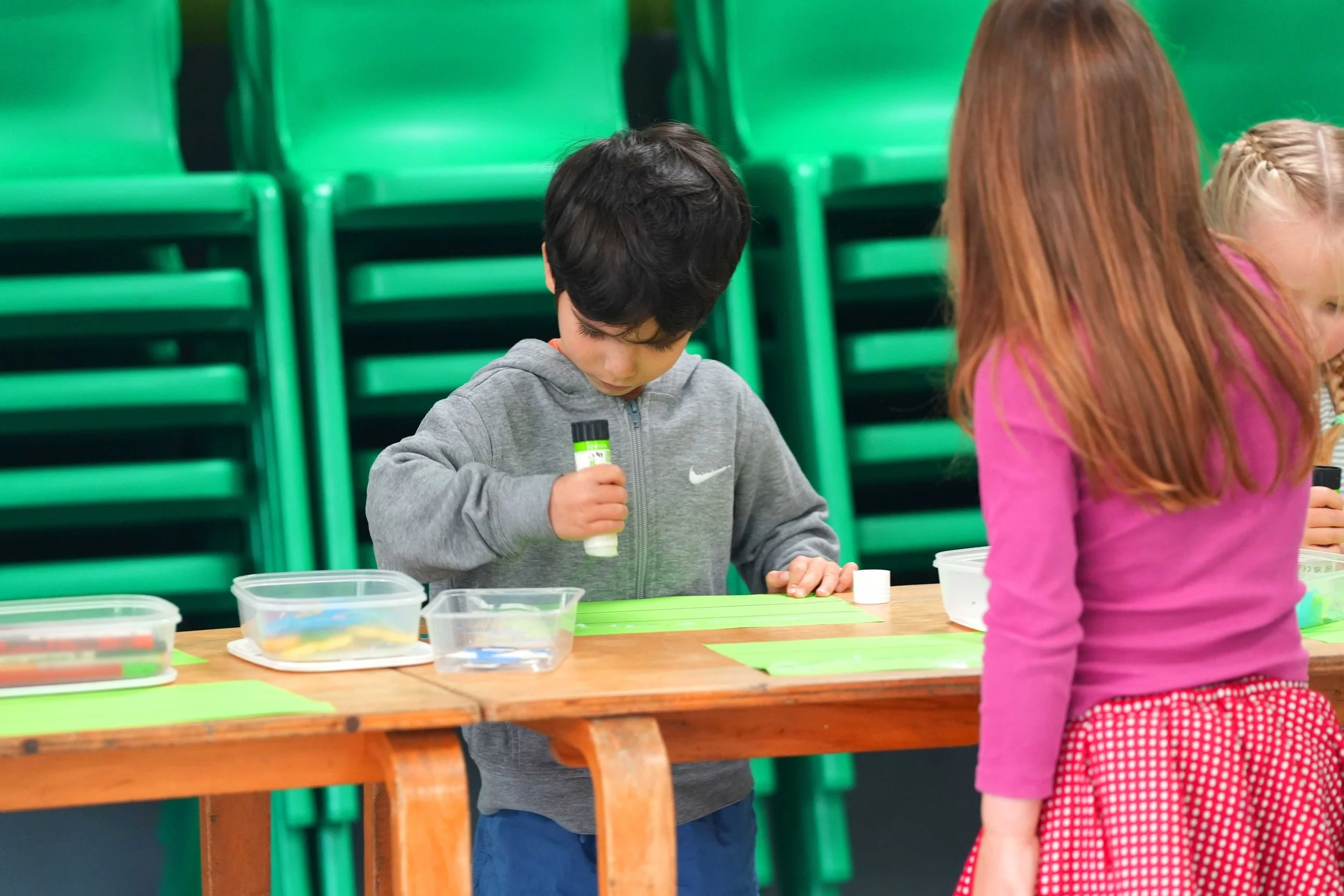 A young boy holding a glue stick, standing at a table with green paper and plastic containers, while a girl with red hair and a pink shirt faces away in the foreground; green chairs are stacked in the background.