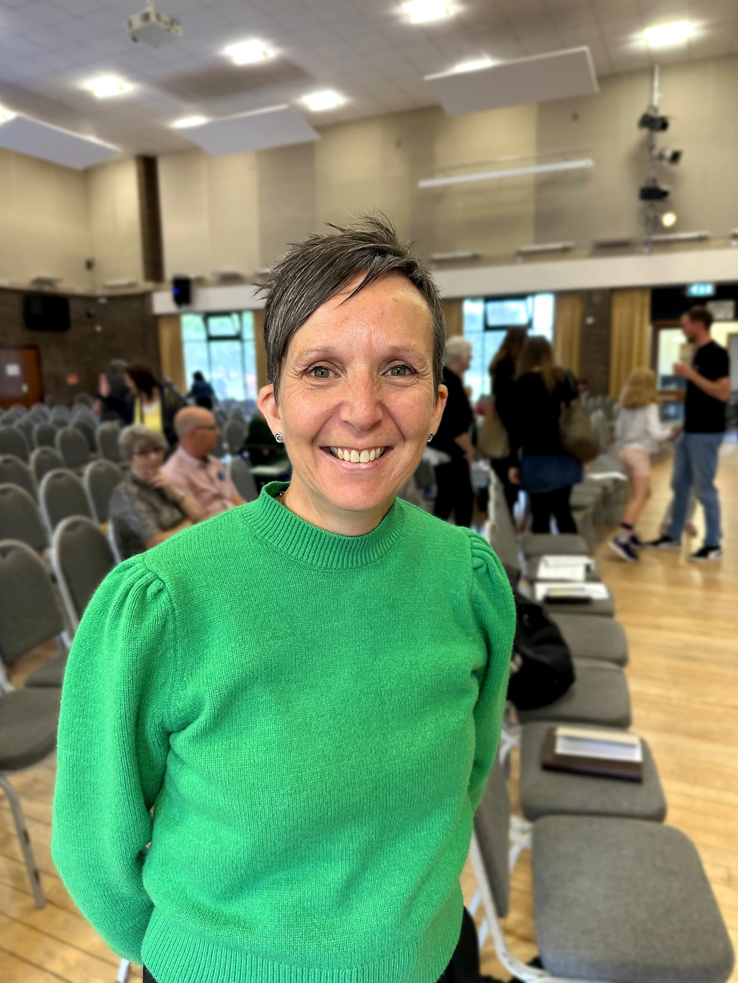 Close-up of a smiling woman with short dark hair wearing a bright green sweater, in a large room with chairs and people in the background.