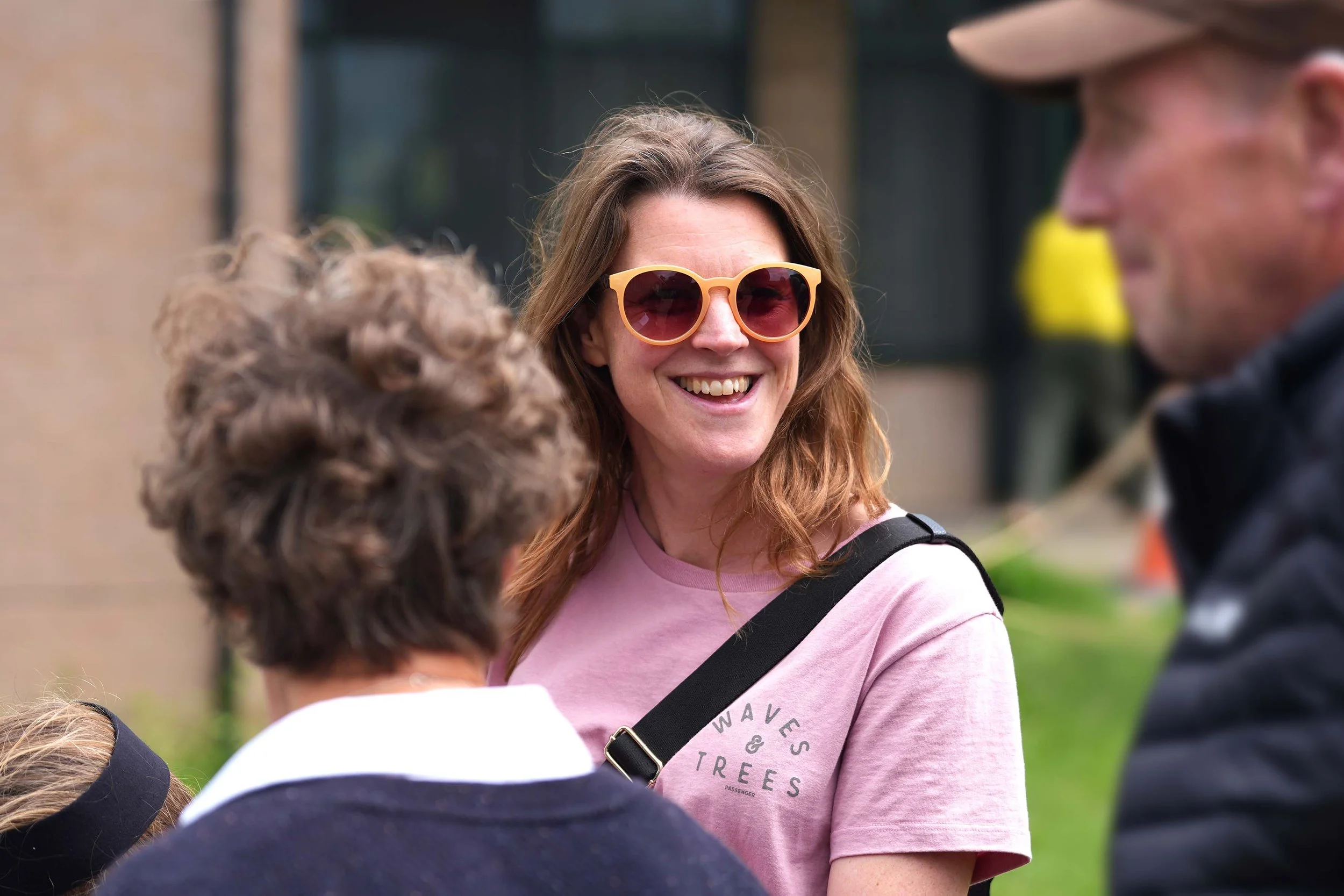 A woman with long brown hair wearing pink sunglasses and a pink t-shirt smiles while talking to children outdoors. Church family at Grace.