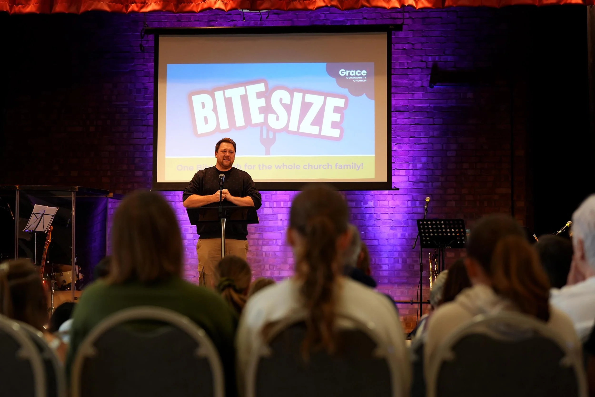 A man standing at a podium on stage in front of a large screen displays a presentation titled 'Bite Size' with the subtitle 'One bite at a time for the whole church family!'. The stage has a brick wall background with purple lighting, and there are musical instruments on the left side of the stage. The audience is seated facing the stage, with some heads visible in the foreground.