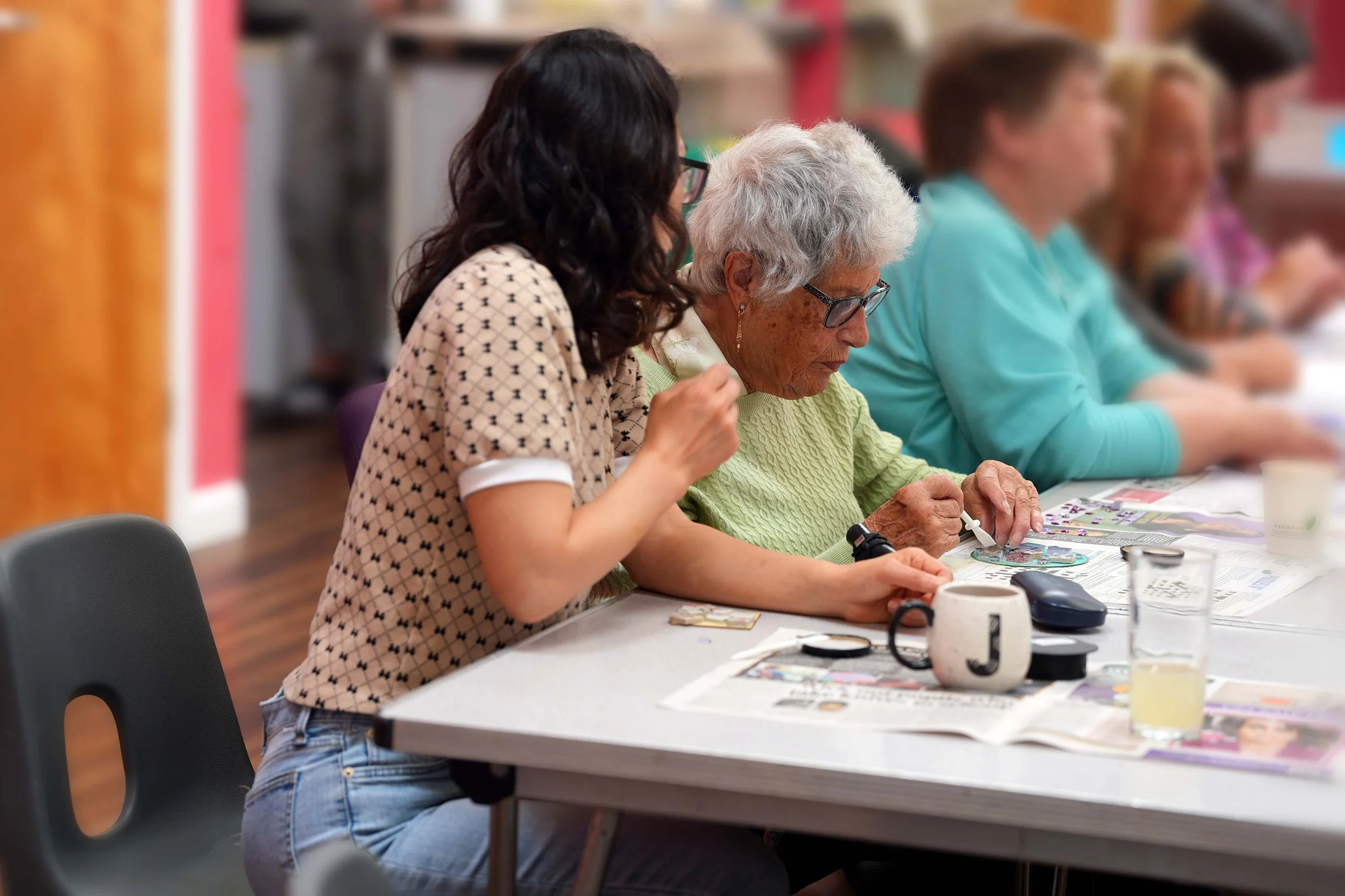 An elderly woman with gray hair and glasses working on a puzzle at a table, with a younger woman assisting her. They are in a communal indoor space with other people in the background.