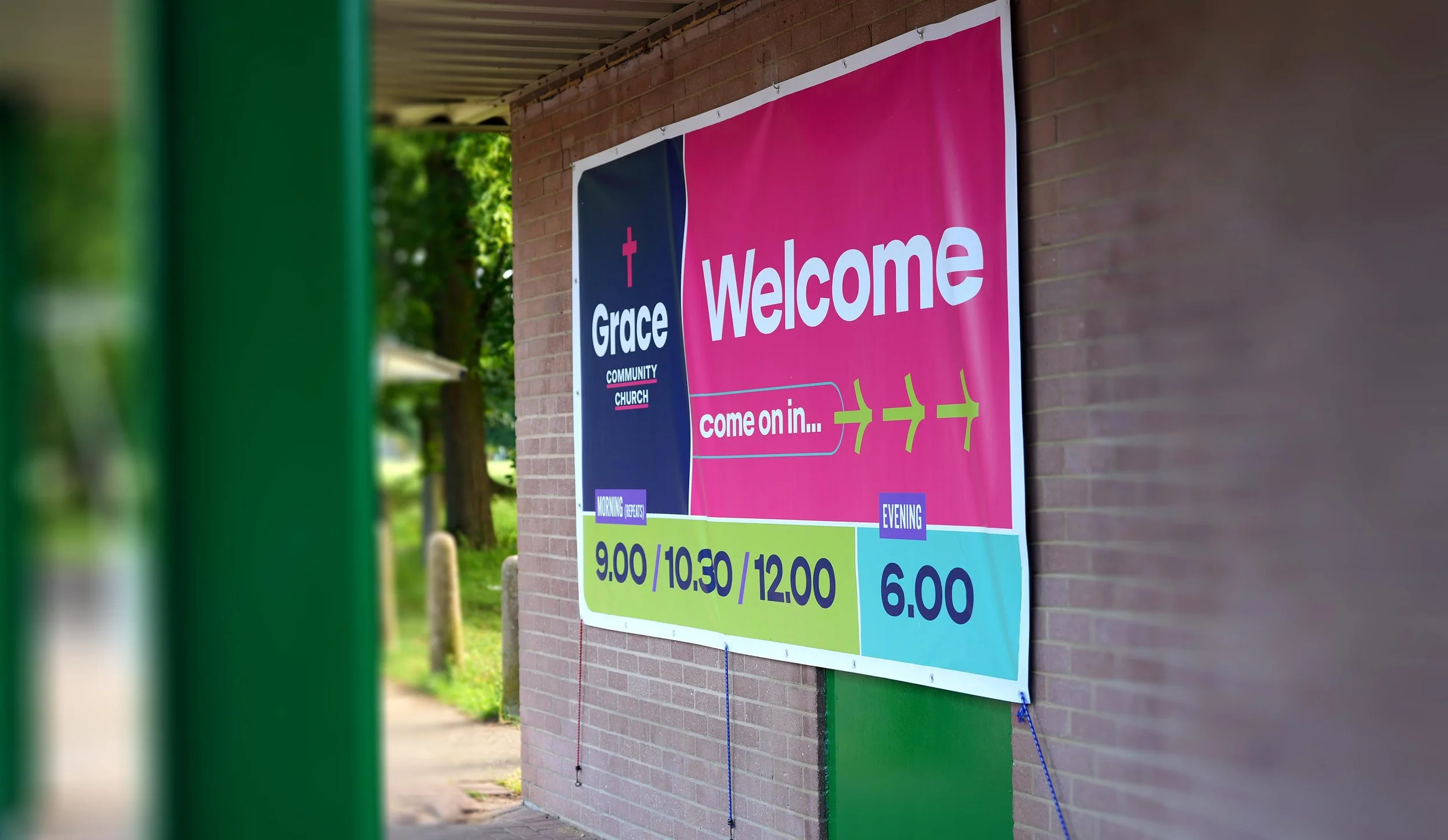 A large colorful welcome sign for Grace Community Church mounted on a brick wall, featuring service times for morning and evening, partially viewed through a green metal frame in the foreground. #Church #SundayServices