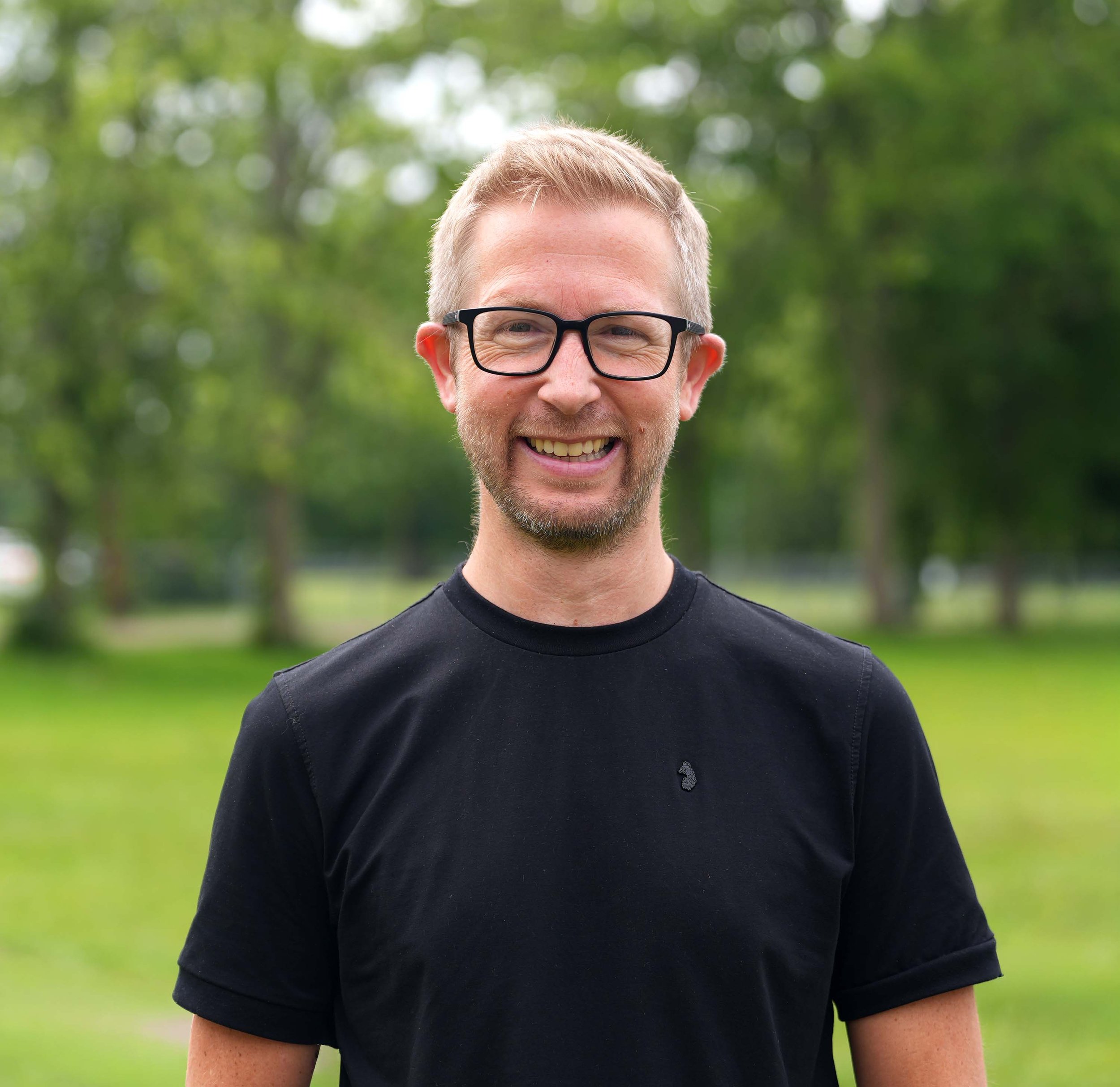 Smiling man with short blond hair, glasses, and a black T-shirt standing outdoors with green trees in the background.