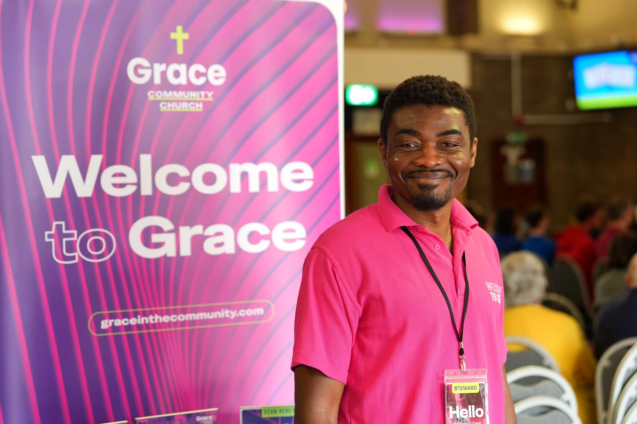 One of the Grace welcome team, standing next to a large purple and pink banner that reads 'Welcome to Grace.' #Church #SundayServices