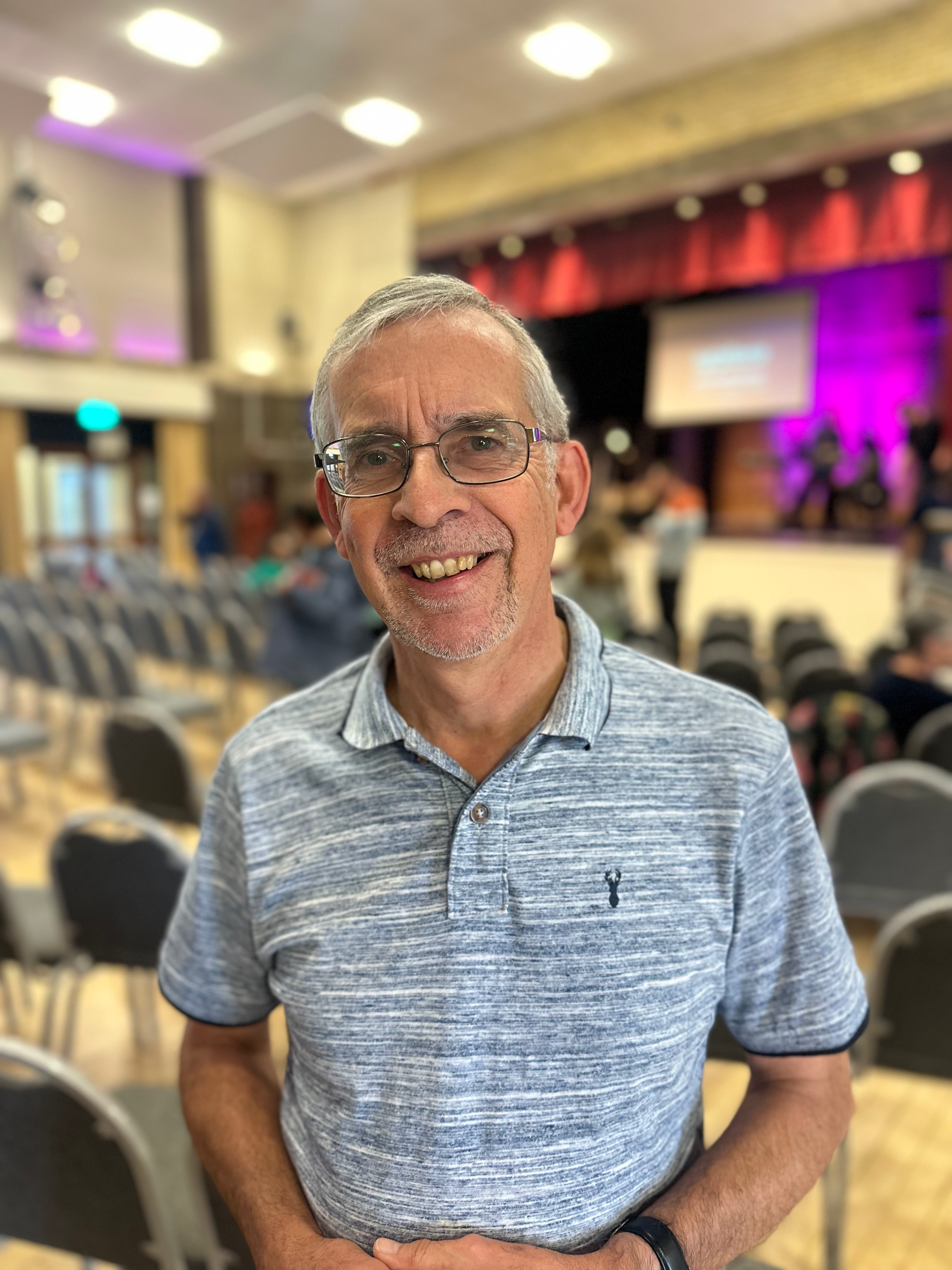 A smiling middle-aged man with glasses and gray hair wearing a gray polo shirt, standing in a large, decorated conference or event room with chairs, screens, and a band on stage in the background.