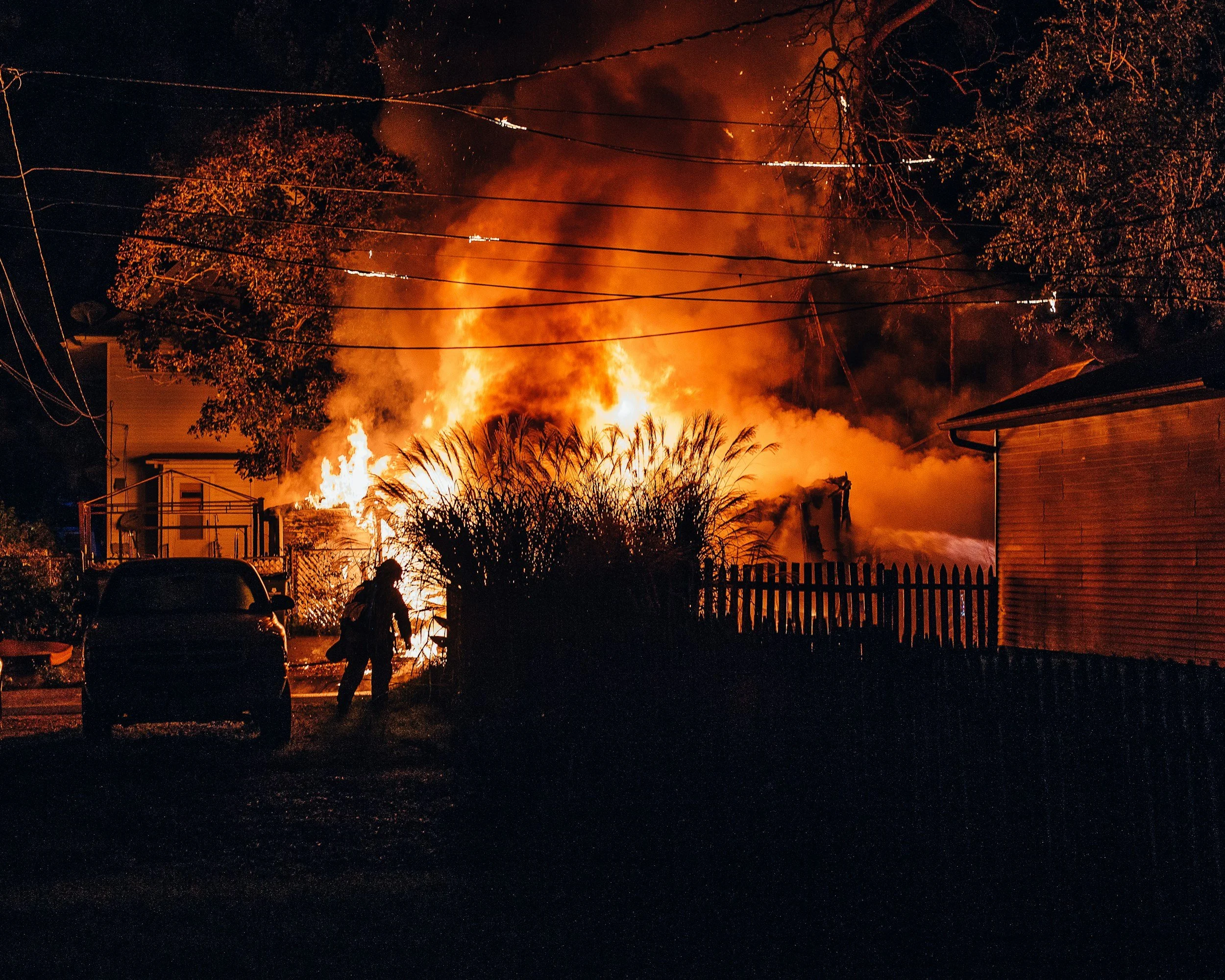 A house on fire at night with large flames and smoke, firefighters and silhouettes of people are visible in the yard, and trees and power lines are overhead.