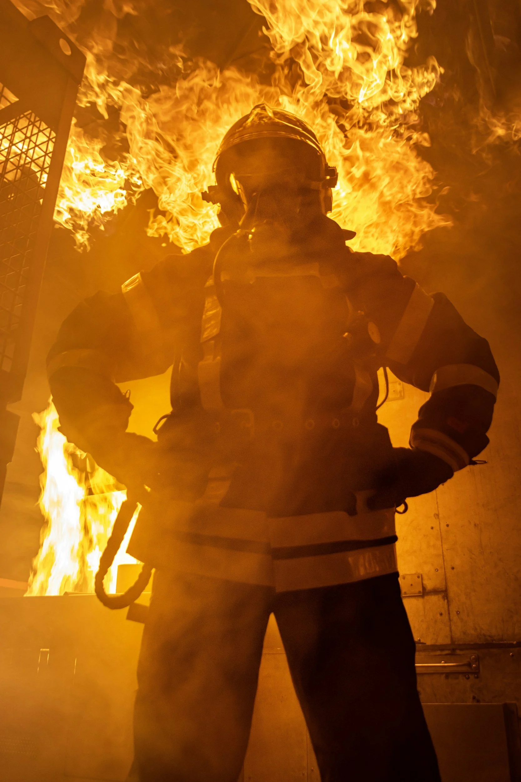 A firefighter wearing protective gear stands in front of flames, with a fire engine visible in the background.