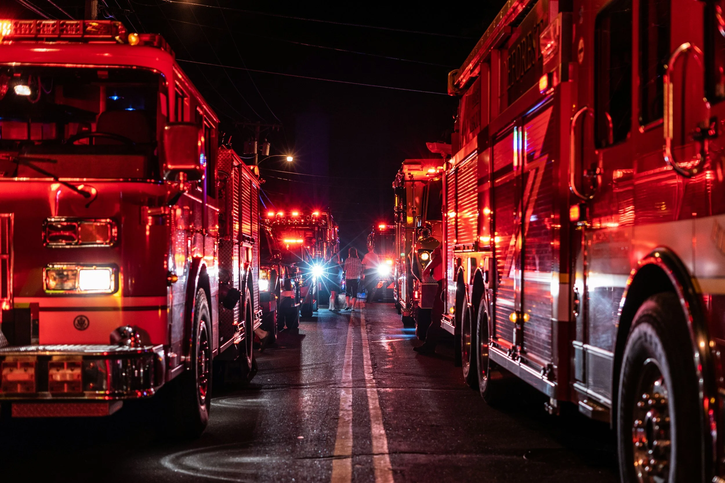 Nighttime scene with multiple fire trucks parked on a wet street, their red emergency lights flashing, and a few people walking between them.