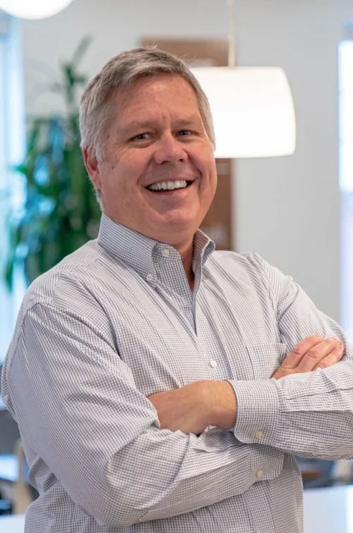 A smiling middle-aged man with gray hair, wearing a light-colored button-up shirt, standing with arms crossed in an office or modern indoor setting.