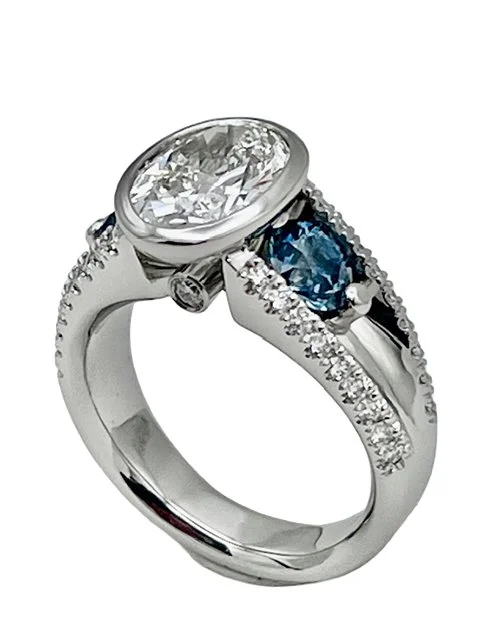 Silver ring with a large oval clear gemstone and a smaller blue gemstone, both surrounded by small diamonds.