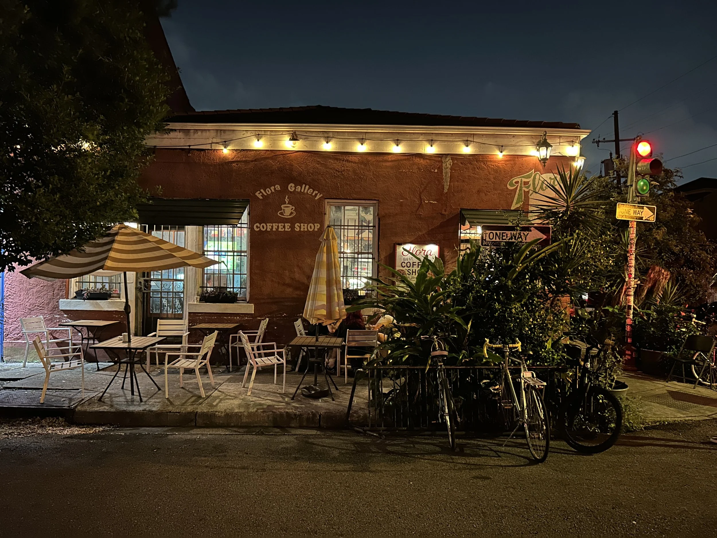 Exterior of Flora Gallery Coffee Shop at night with string lights, patio furniture, bicycles, and a traffic light showing red.
