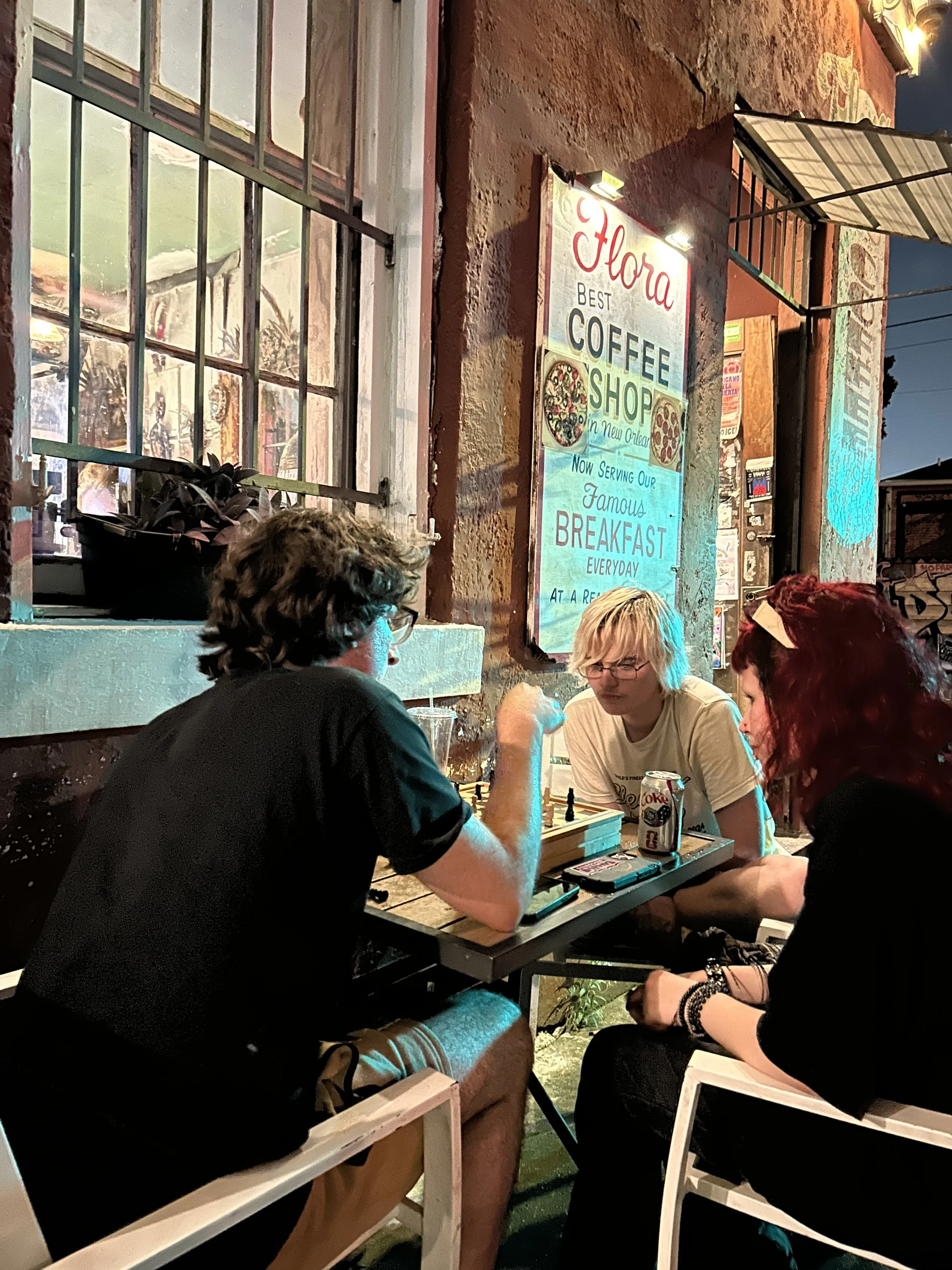 People playing chess at an outdoor evening gathering with a rustic building featuring a sign for a coffee shop in the background.