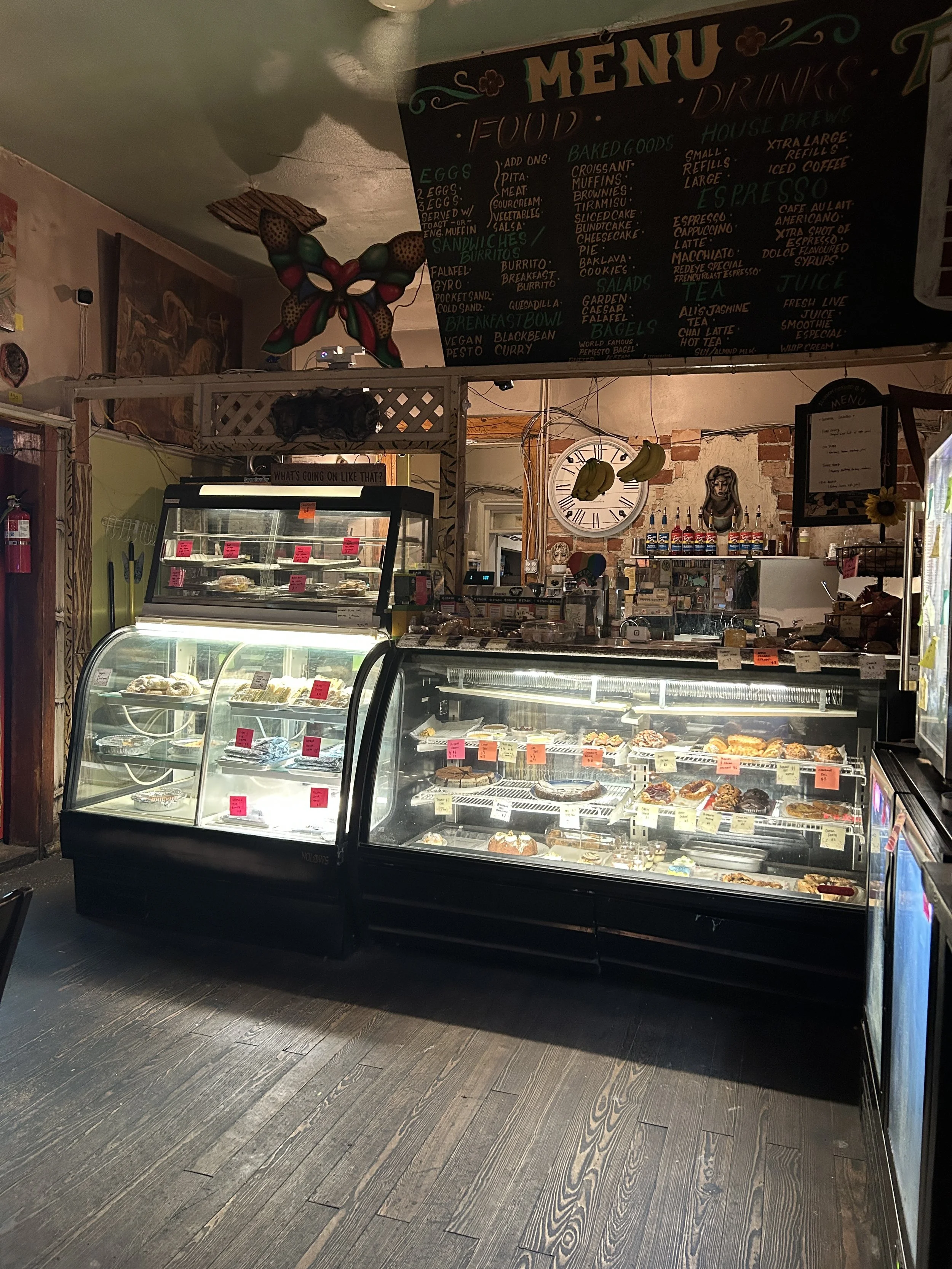Inside a bakery or cafe with a display case filled with various pastries and baked goods. There is a black menu board hanging above with handwritten items, a decorative butterfly sculpture on the ceiling, a clock, bananas hanging, and various decorations and supplies on the counter and walls.