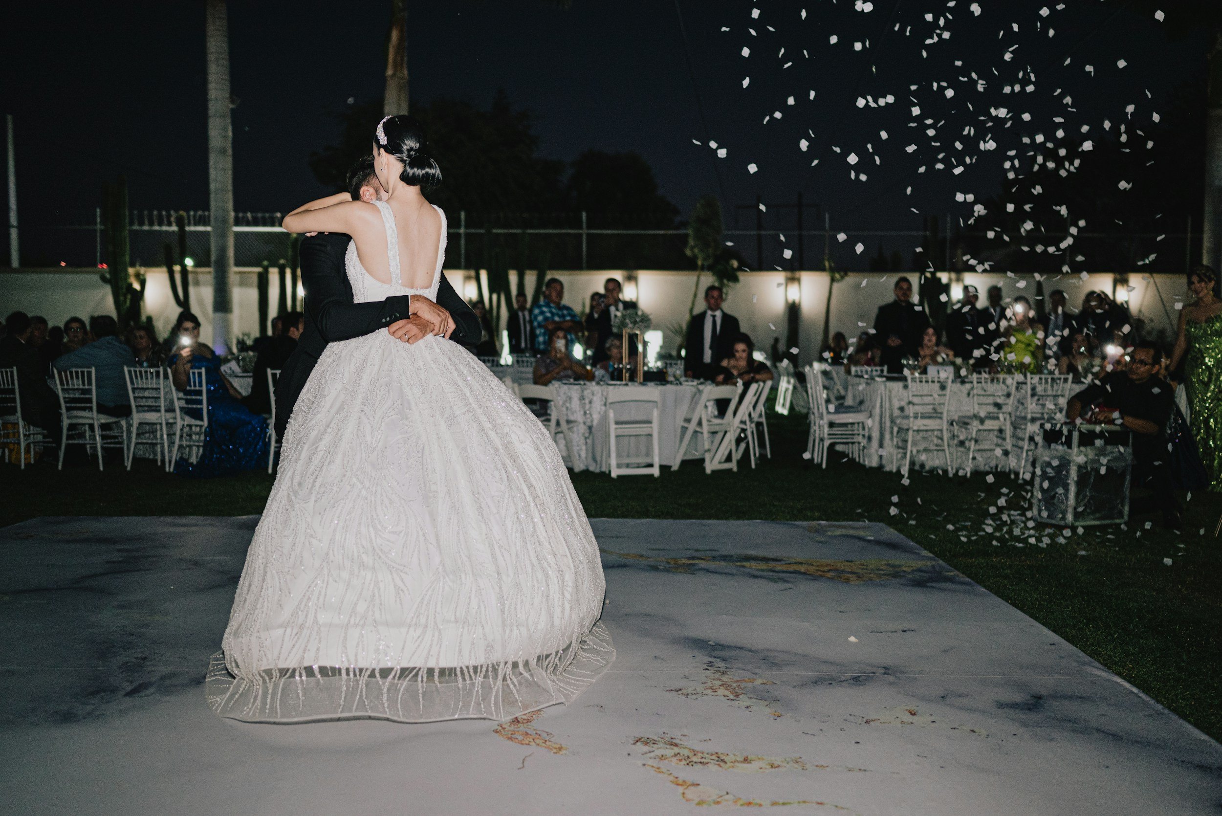 A newlywed couple shares a dance at their wedding reception during nighttime, with guests seated at decorated tables in the background and white confetti falling from above.