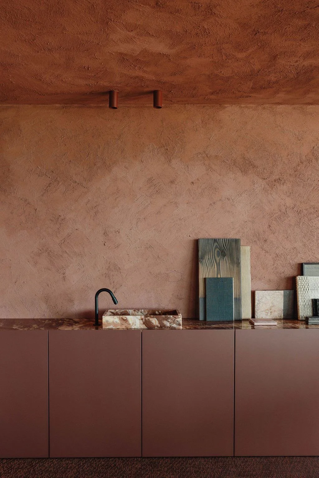 Parete rosa con credenza di colore marrone e decorazioni in legno e pietra, con sedia di tessuto chiaro e elementi decorativi su superficie.
