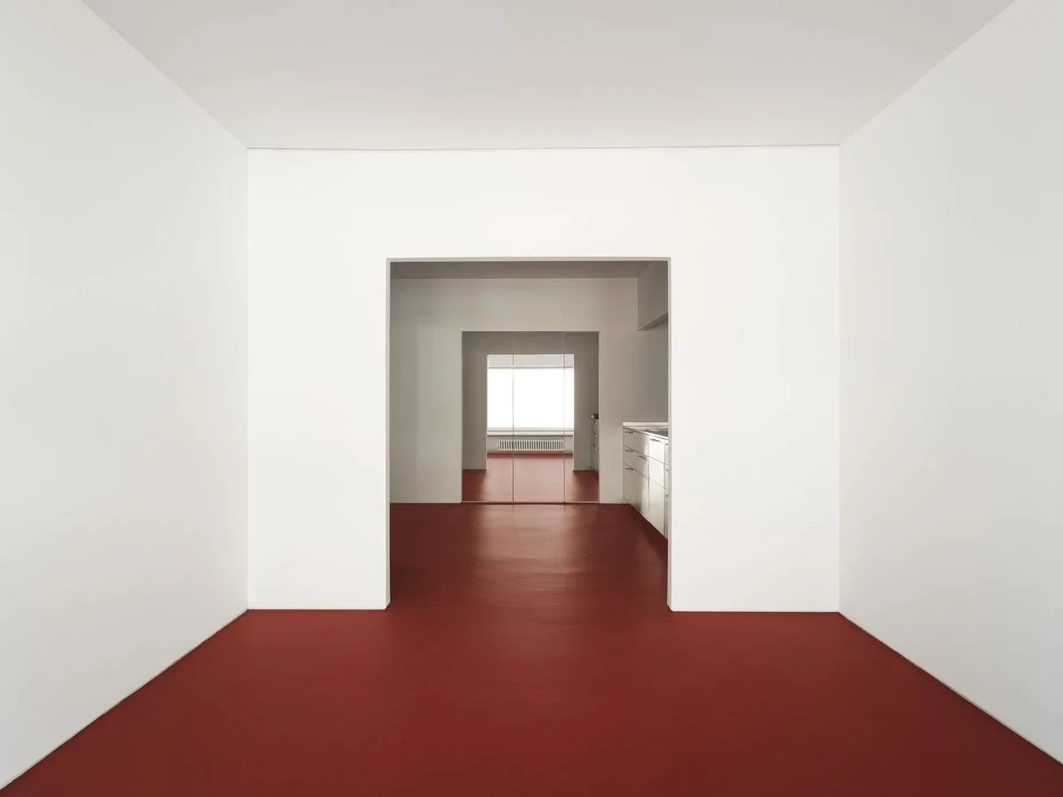 Interno di un appartamento con pareti bianche e pavimento rosso, vista verso una stanza con finestra.