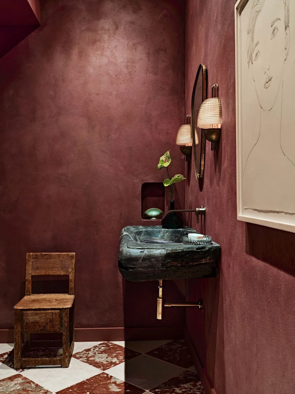 Interno con pareti rosse, sedia di legno, lavabo in pietra con una pianta verde, due specchi rotondi e un quadro con ritratto femminile minimalista