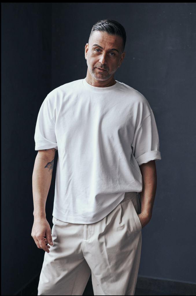 Uomo con capelli corti e barba, indossa una maglietta bianca e pantaloni beige, posa contro uno sfondo scuro.