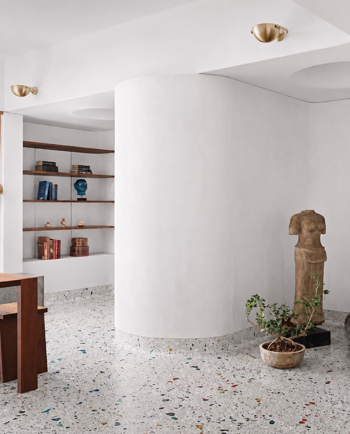 Interno di una casa con pareti bianche, scaffali con libri e oggetti decorativi, statua femminile in pietra, pianta in vaso e pavimento in terrazzo colorato.