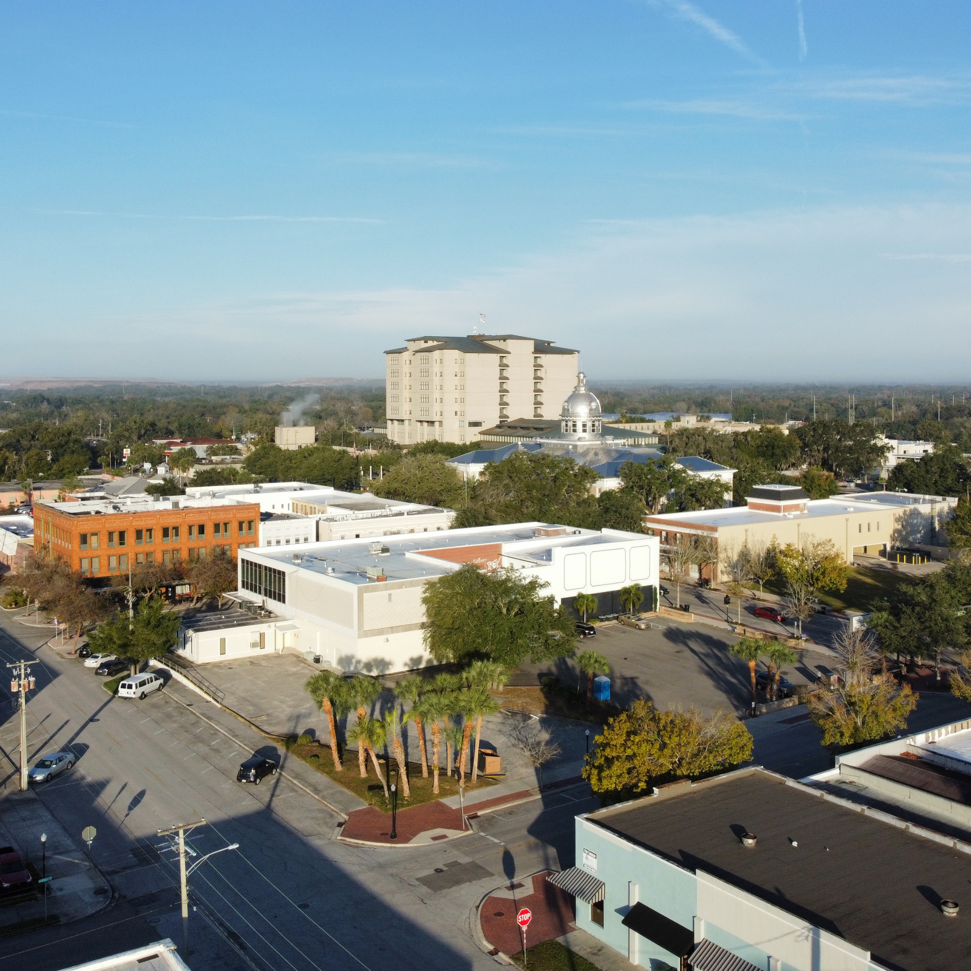 A cityscape with a tall cream-colored building, a smaller domed structure, and palm trees in the foreground, under a blue sky with wispy clouds.