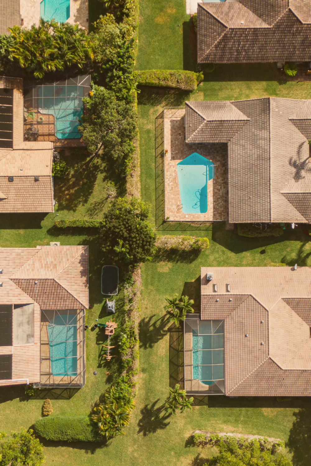 Aerial view of suburban houses with swimming pools, some enclosed in screened enclosures, surrounded by green lawns, trees, and shrubs.