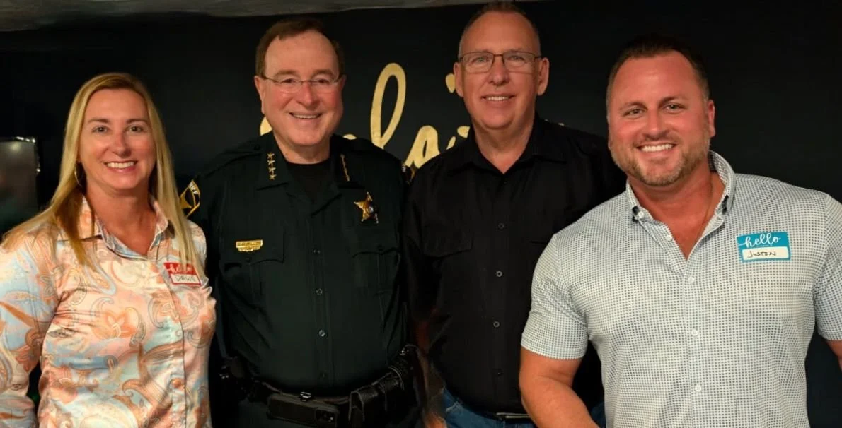 Four people standing together, smiling at the camera, in front of a black backdrop with gold writing. The group includes a woman on the left, a police officer in uniform, a man with glasses, and a man wearing a name tag that says "Justin" on the right.