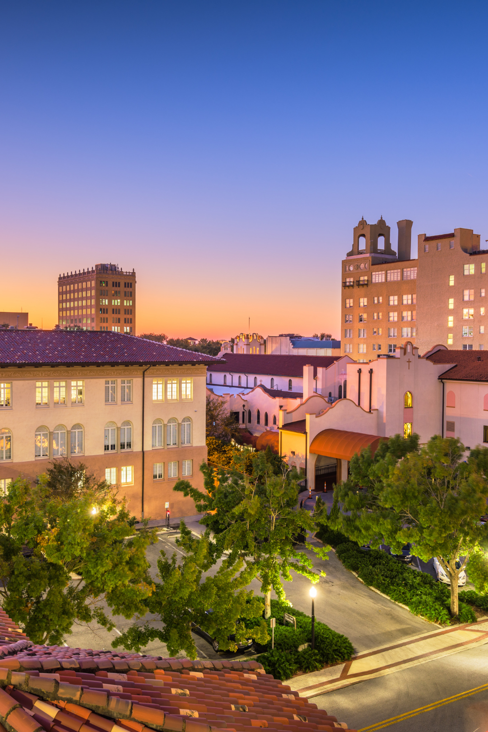 Cityscape at sunset with colorful sky, trees, and buildings including a church-like structure and taller modern buildings in the background.