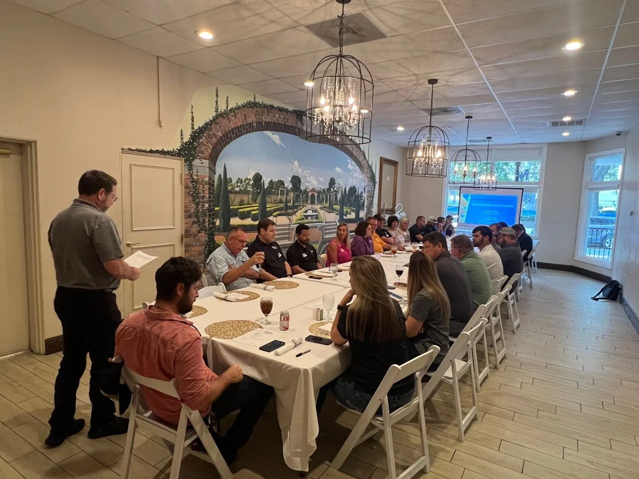 People seated around a long conference table in a restaurant, attending a meeting or presentation. A man is standing and reading from a paper, while a large screen displays a presentation at the end of the room. The room has hanging chandeliers and a mural of a European garden on the wall.