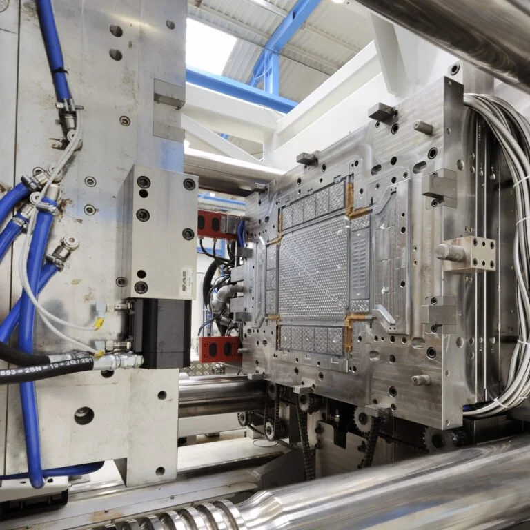 Close-up of a metal injection mold with hydraulic and pneumatic tubing, mounted on a machine in a manufacturing facility.