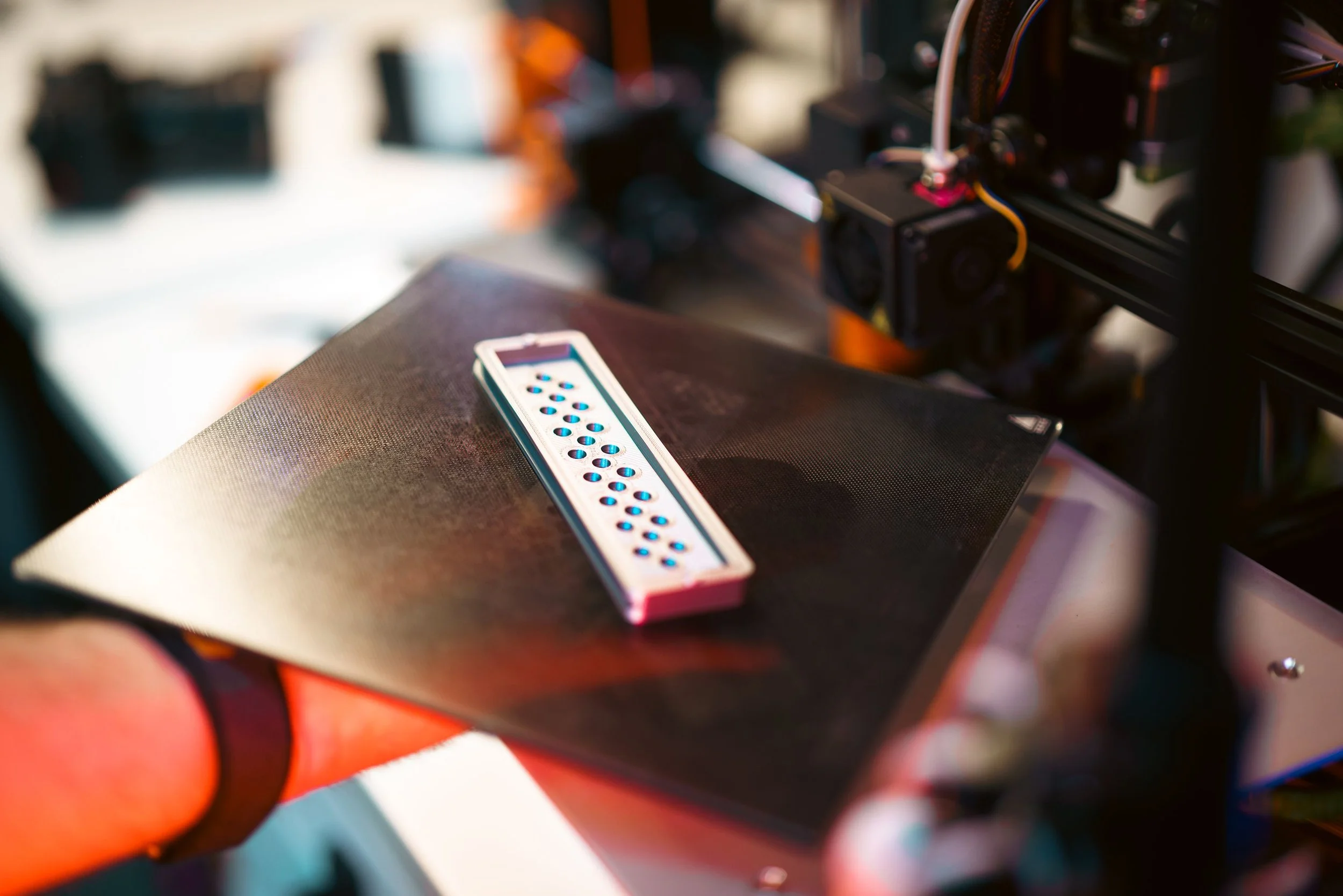 Close-up of a 3D printer with a black and orange frame, with a white and pink filament holder resting on a black surface in the foreground.