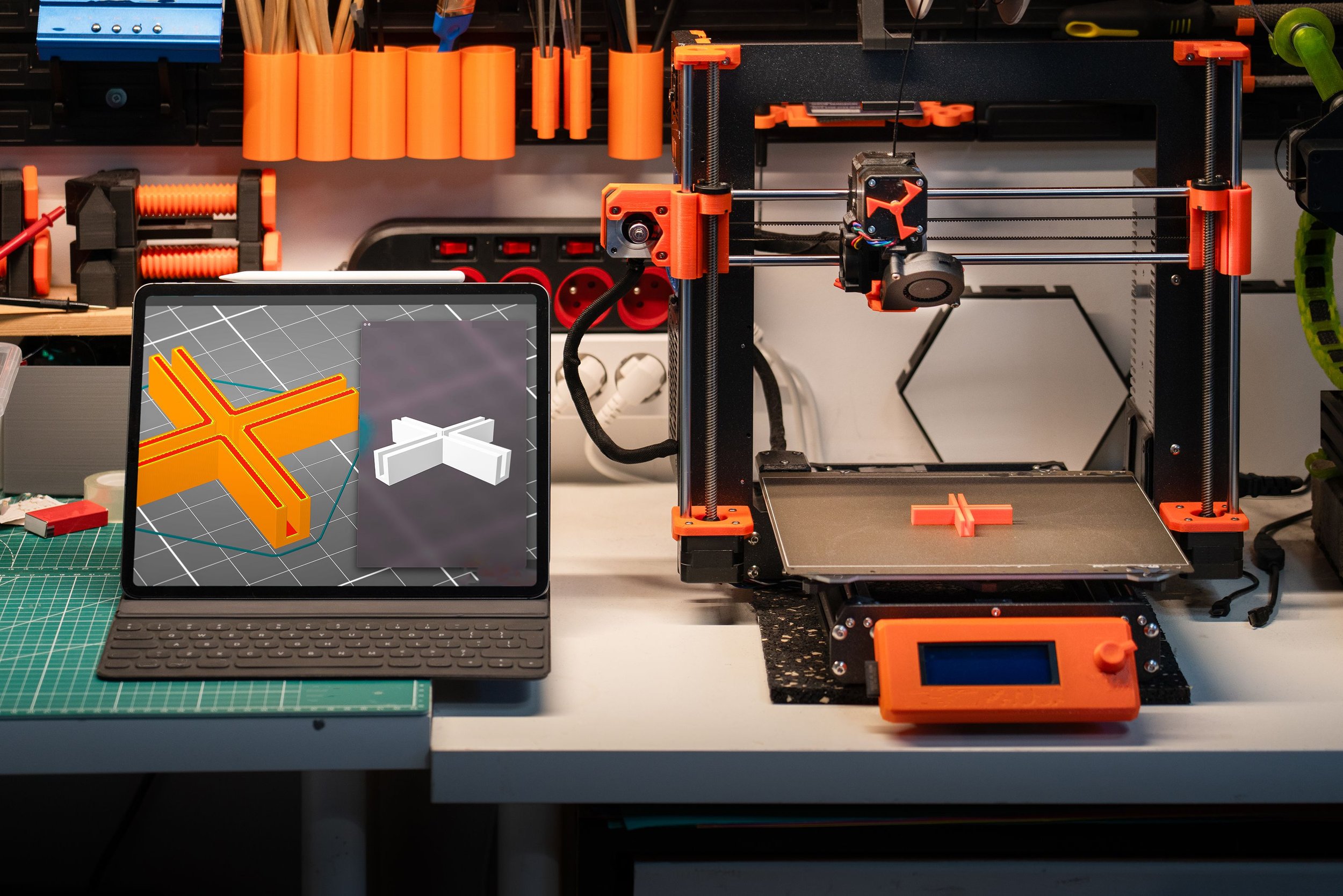 A workspace with a 3D printer, a laptop displaying a 3D model of an orange T-shaped object, and various tools and materials around, including orange filament spools and green cutting mats.