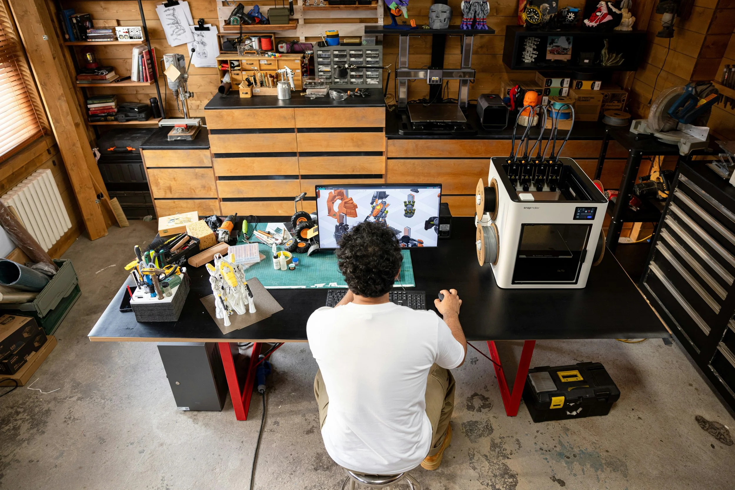 A person working at a cluttered craft or maker space table with a computer, 3D printer, and various tools and models. The room has wooden walls, shelves with books and supplies, and a window with blinds.