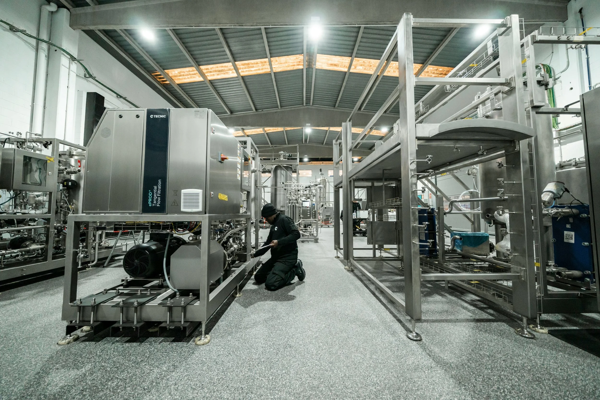 Industrial facility with stainless steel machinery and pipes, worker kneeling in front of equipment, high ceiling with skylights.