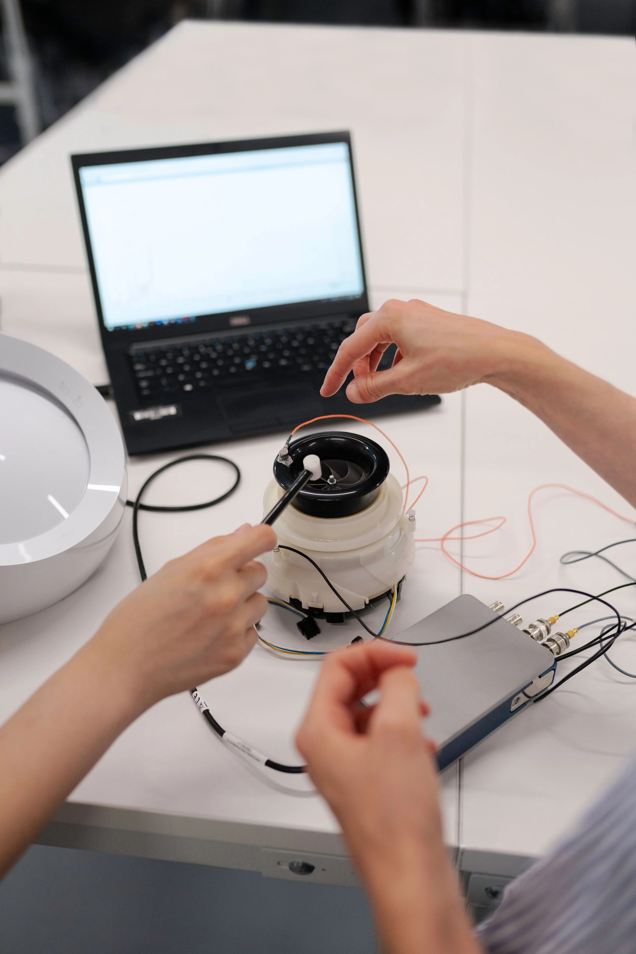 Hands working on a scientific experiment with wires and equipment on a white table, a laptop displaying data in the background.