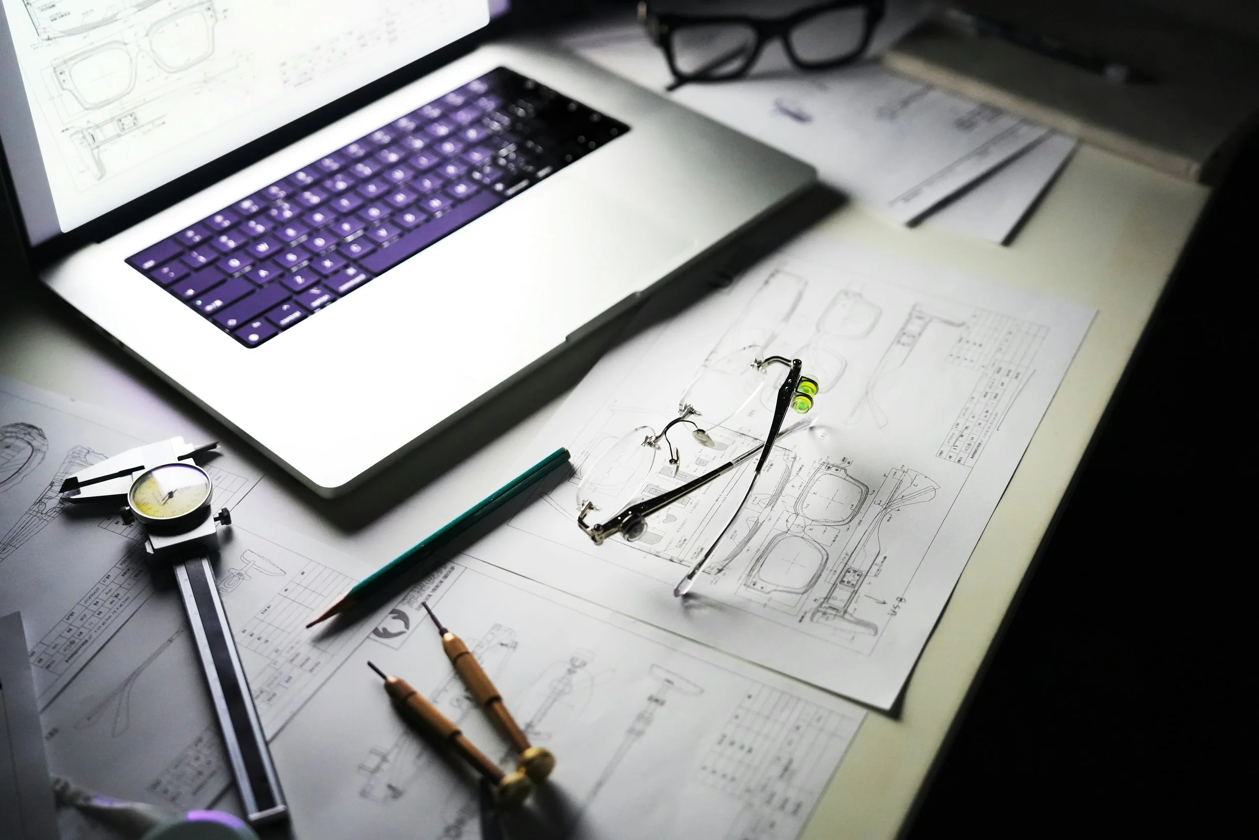 Desk with technical drawings, a pair of safety glasses, a green pencil, and a mechanical stopwatch, along with a laptop and eyeglasses in the background.