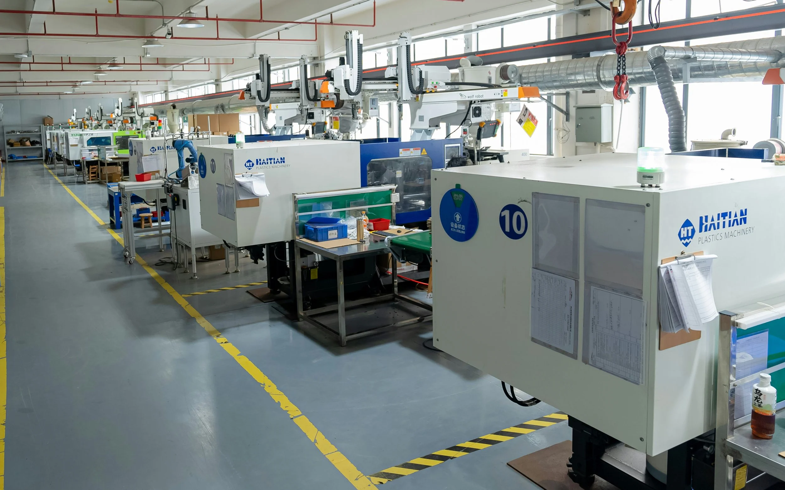 Factory or industrial workspace with machinery, workbenches, and equipment organized in rows, including several large white machines with blue labels, and overhead pipes.