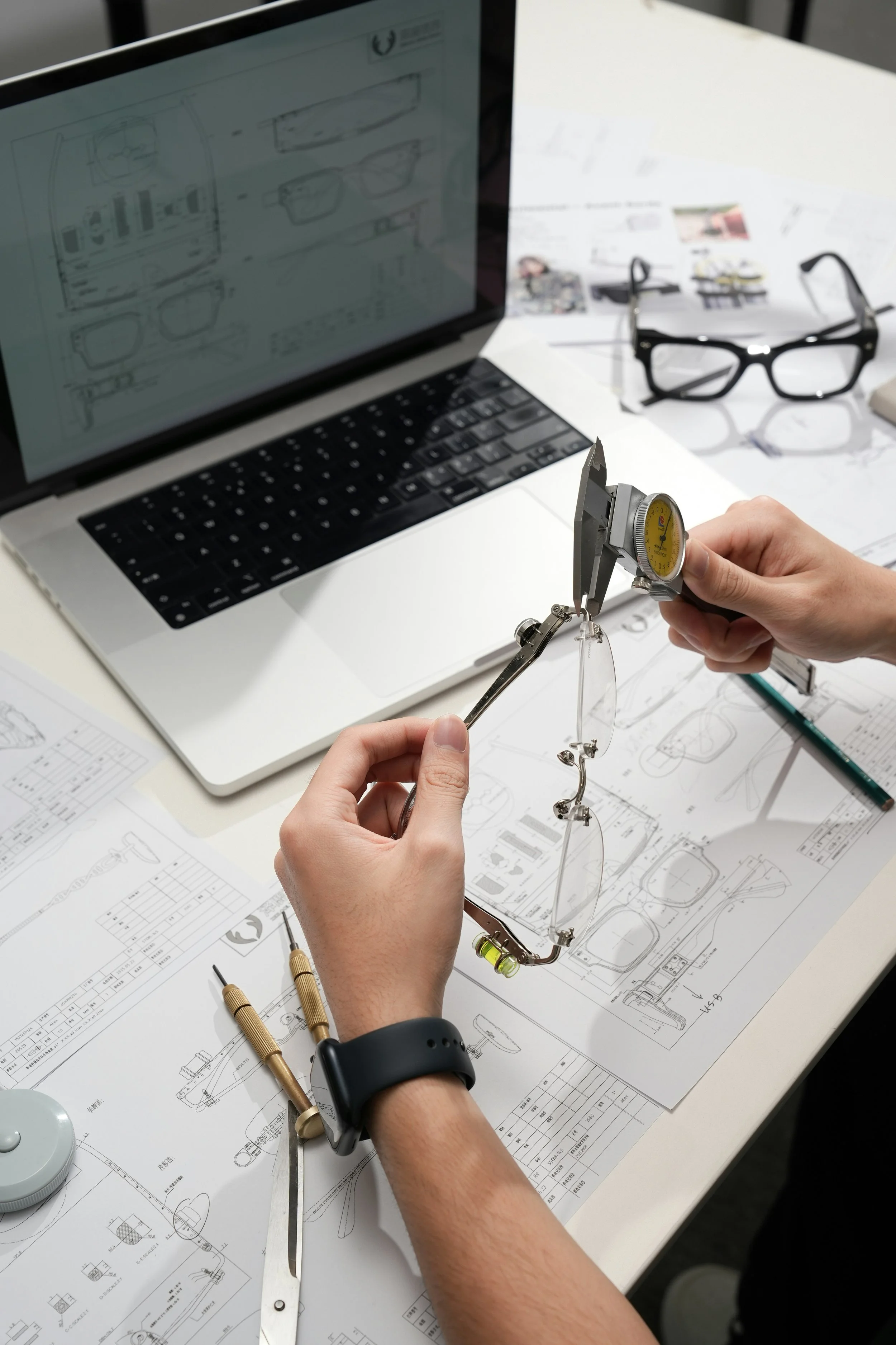 A person working on eyeglass fitting with a caliper, surrounded by technical diagrams and tools on a white table, with a laptop and additional eyeglasses in the background.