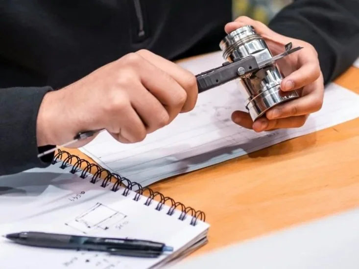 Person examining a microscope slide with a small metal object, sitting at a wooden table with notebook and pen.