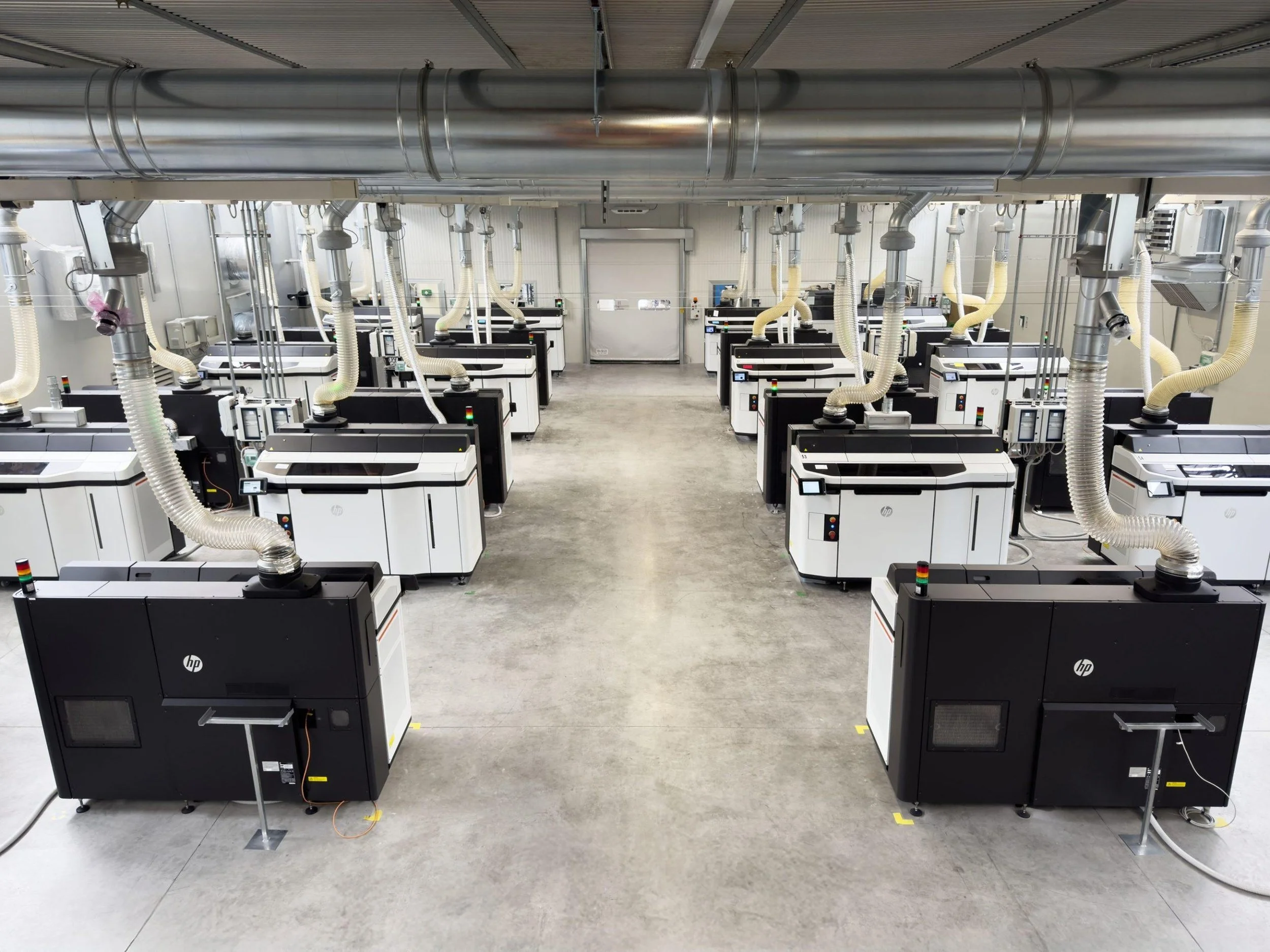 A large industrial room filled with multiple 3D printers positioned on the floor in rows, connected to numerous tubes and cables, with a metallic ductwork ceiling overhead.