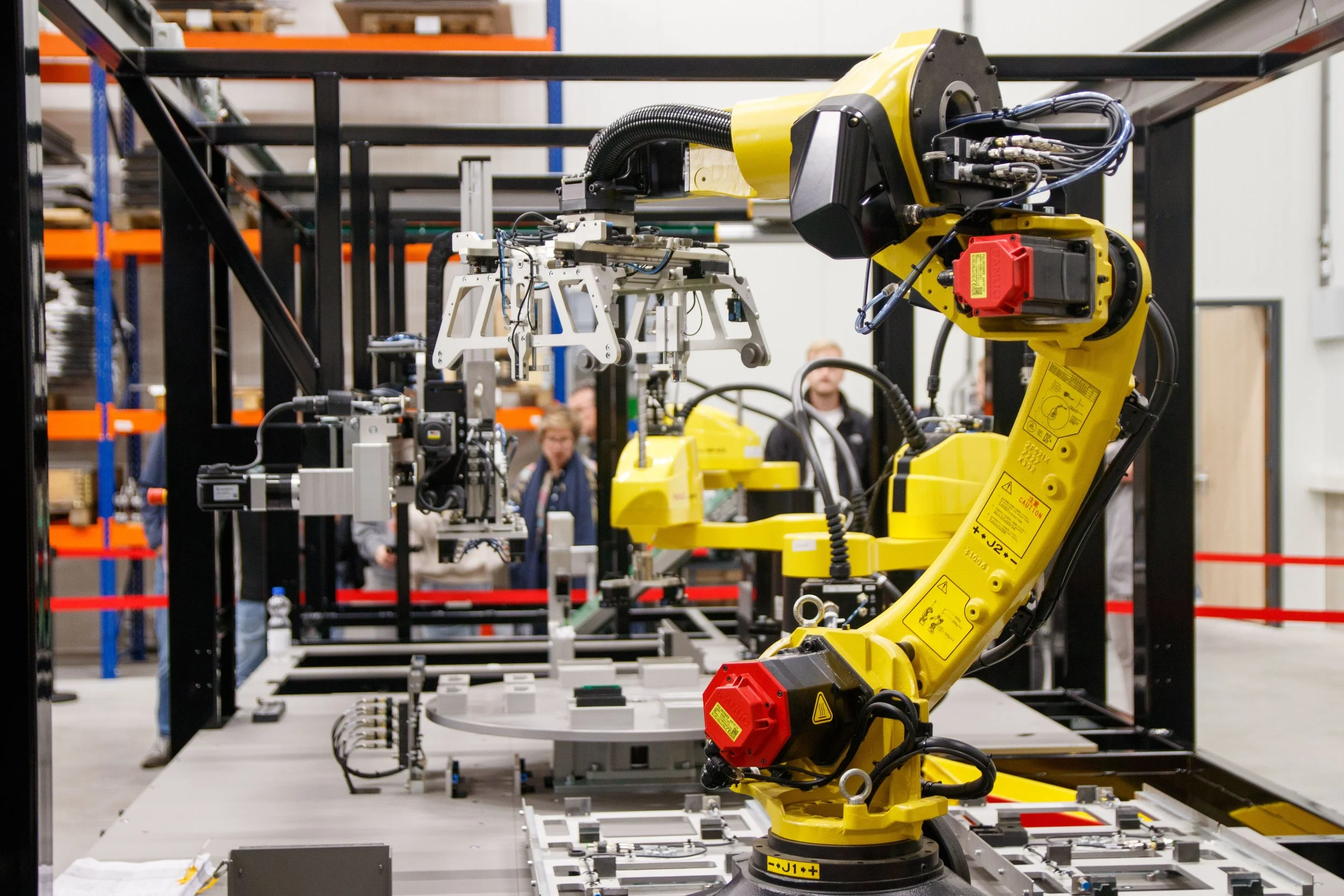 Industrial robotic arm working on machinery in a factory environment with people observing in the background.
