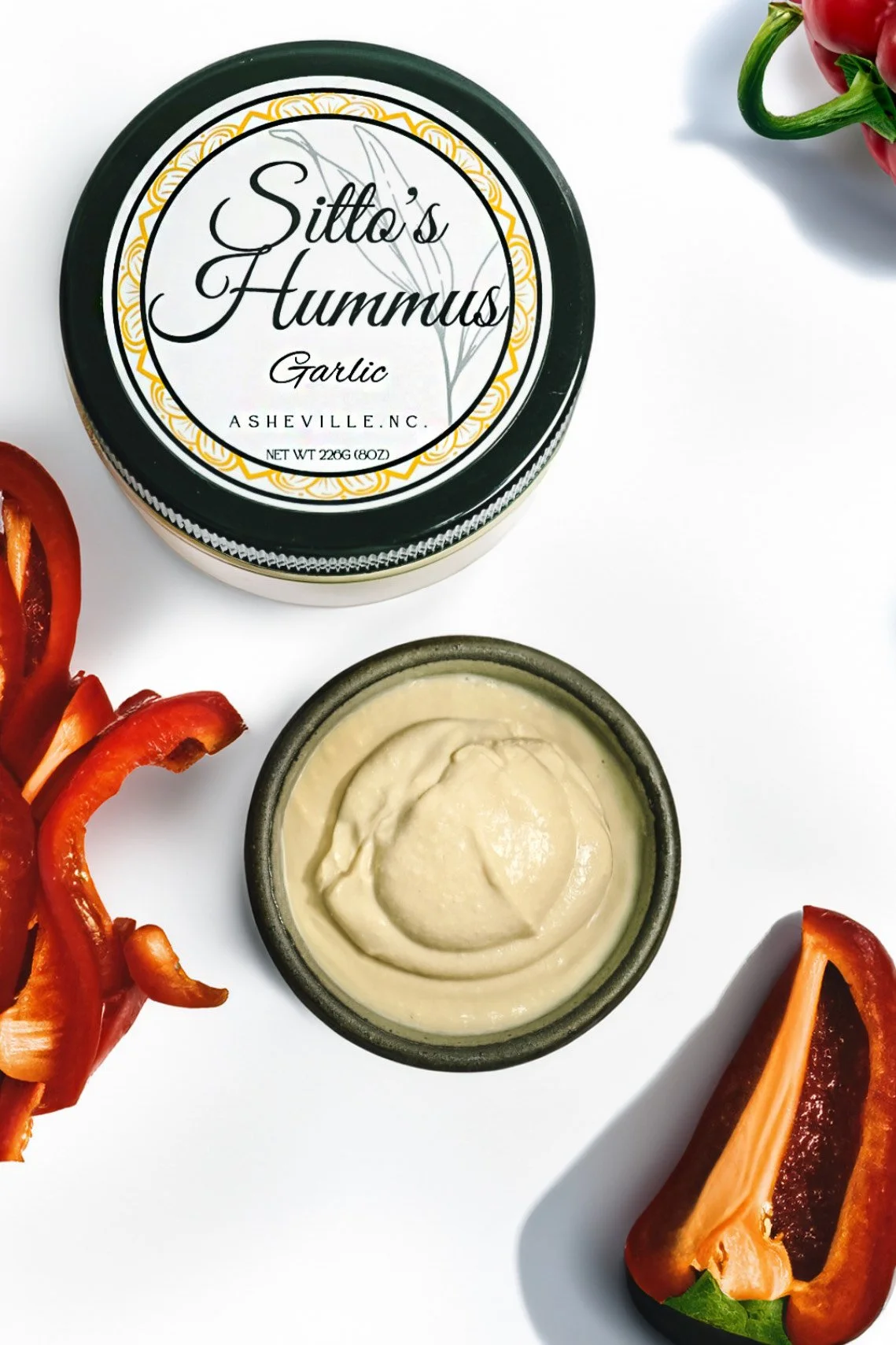 Container of Sittlo's Hummus Garlic with a small bowl of hummus, red bell pepper slices, and a red bell pepper on a white surface.