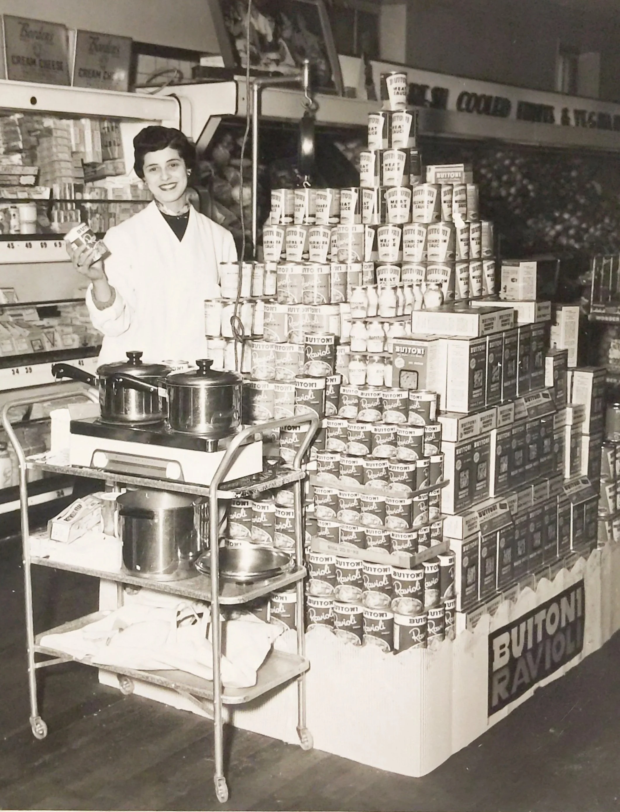 A woman in a white coat is standing behind a cart with pots and a box, holding a jar in her right hand. She is at a grocery store aisle, with shelves stocked with canned goods and boxes, and a sign reading 'Canned Goods & Tins' in the background.
